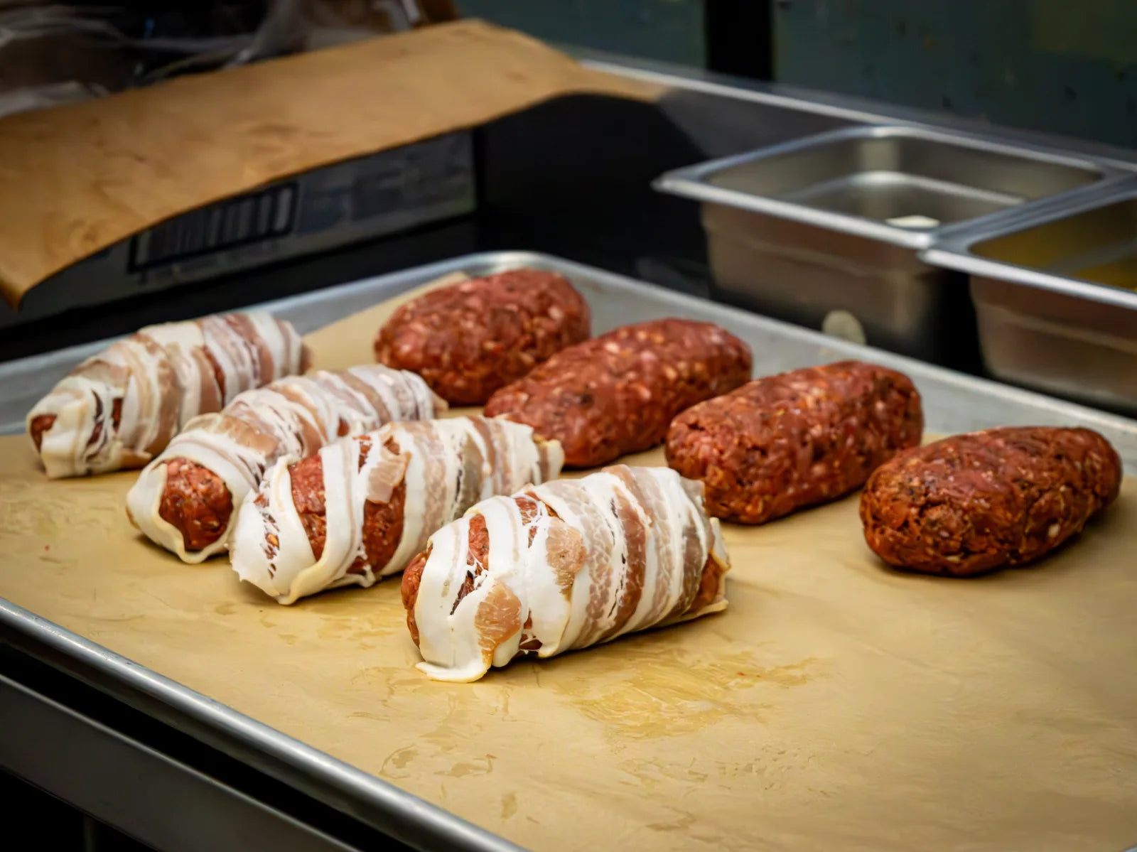 Six raw meat rolls on a metal prep tray, three wrapped in woven strips of bacon and three coated in dark seasoning.