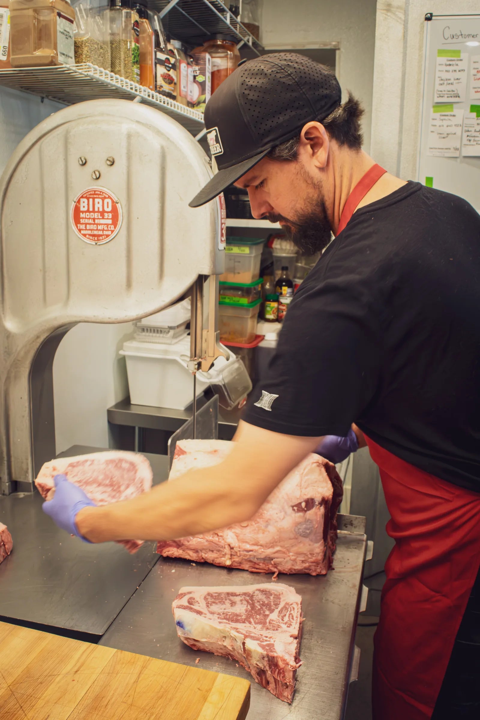 Butcher in black cap and red apron pulling a cut steak away from a band saw, with a portioned bone-in steak on the table.