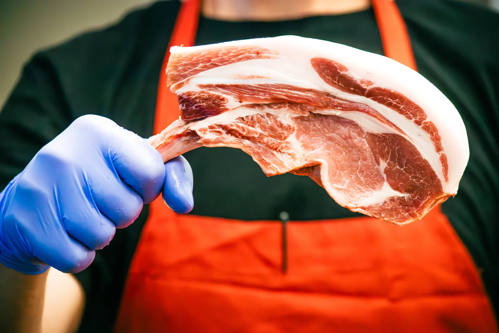 Gloved hand holding up a raw bone-in pork chop with visible marbling, framed against a butcher's dark shirt and red apron.
