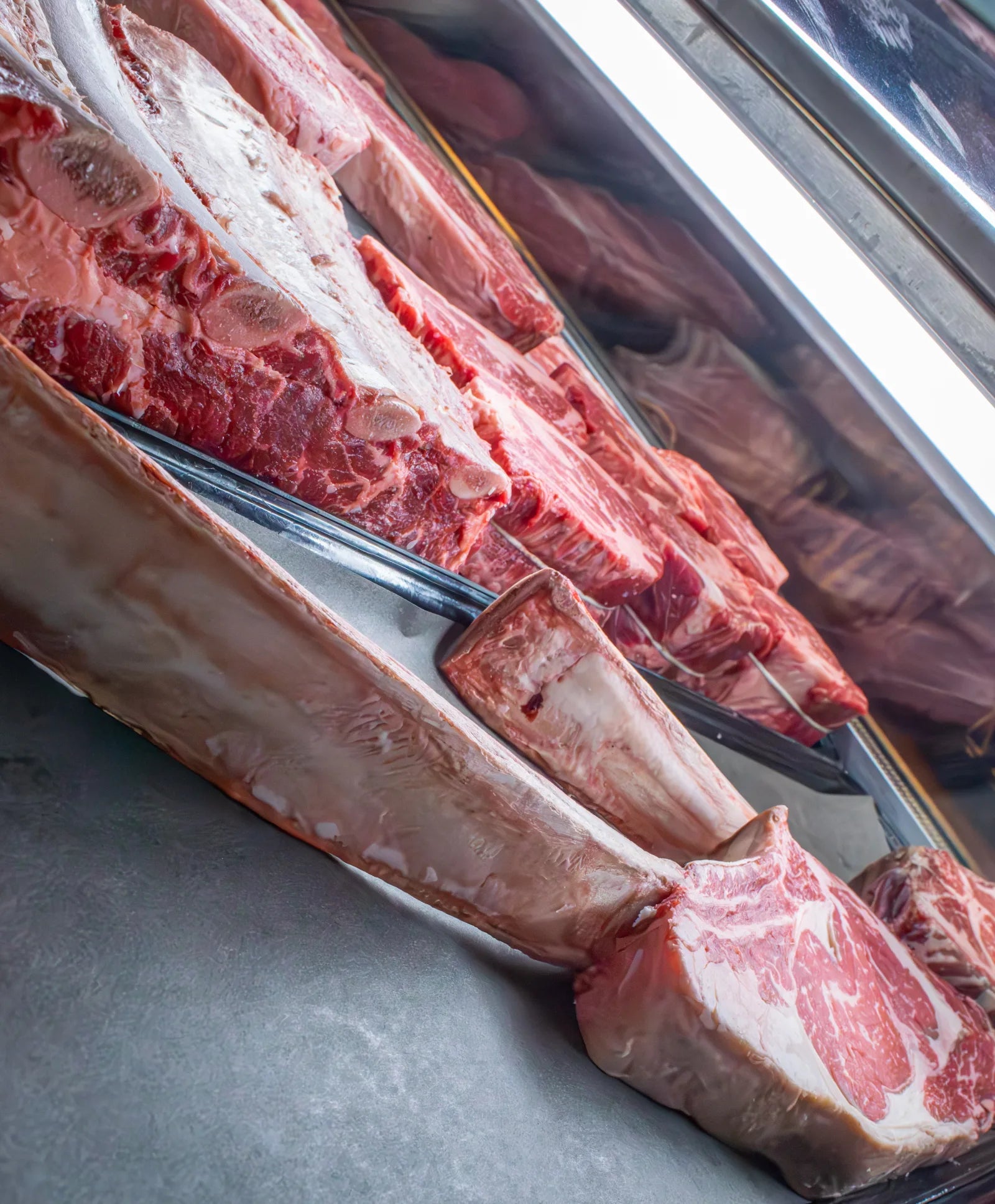 Row of raw beef primal cuts hanging on a stainless meat rail with hooks, backlit by bright window light.