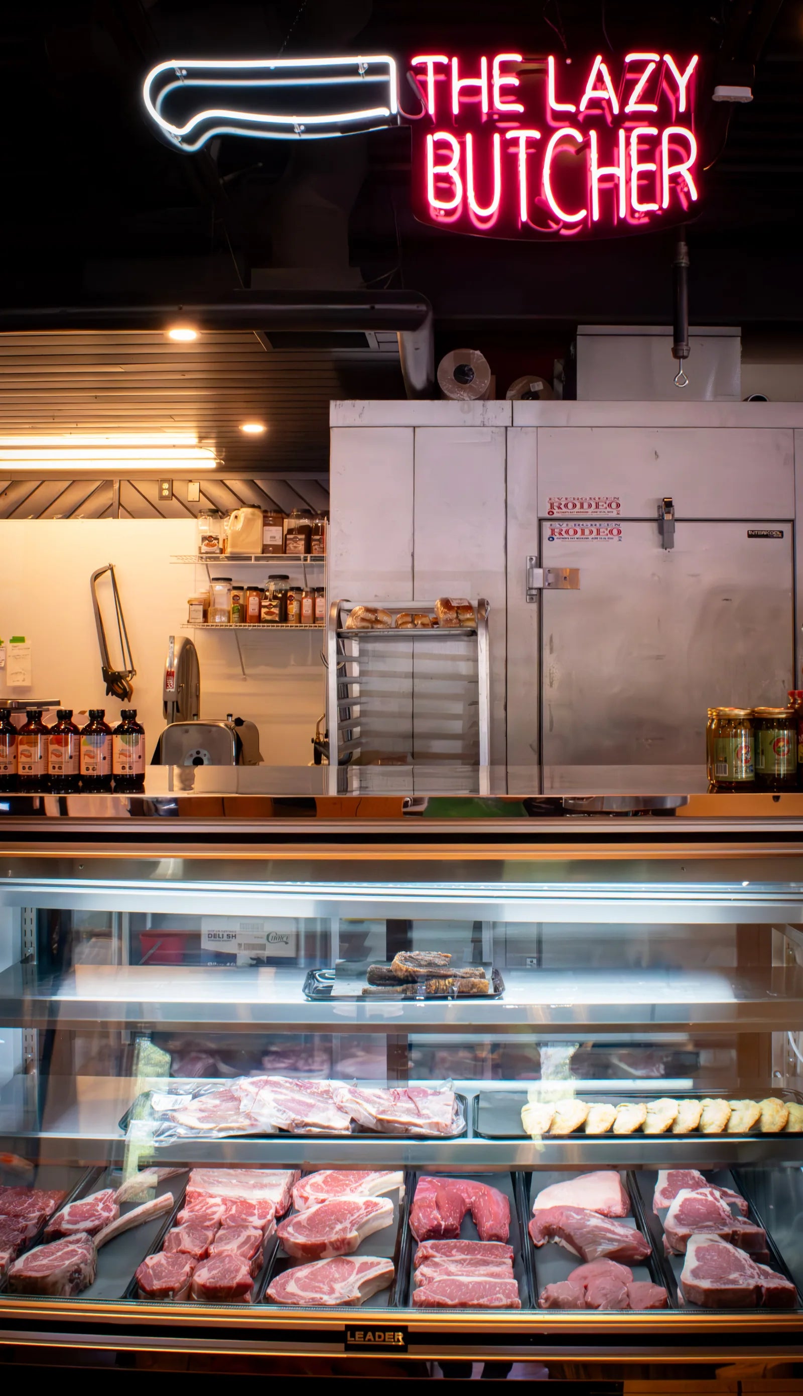 Glowing neon sign reading 'The Lazy Butcher' with a knife icon, mounted above a glass butcher case filled with rows of raw steaks, a walk-in cooler, and shelved jars in the background.