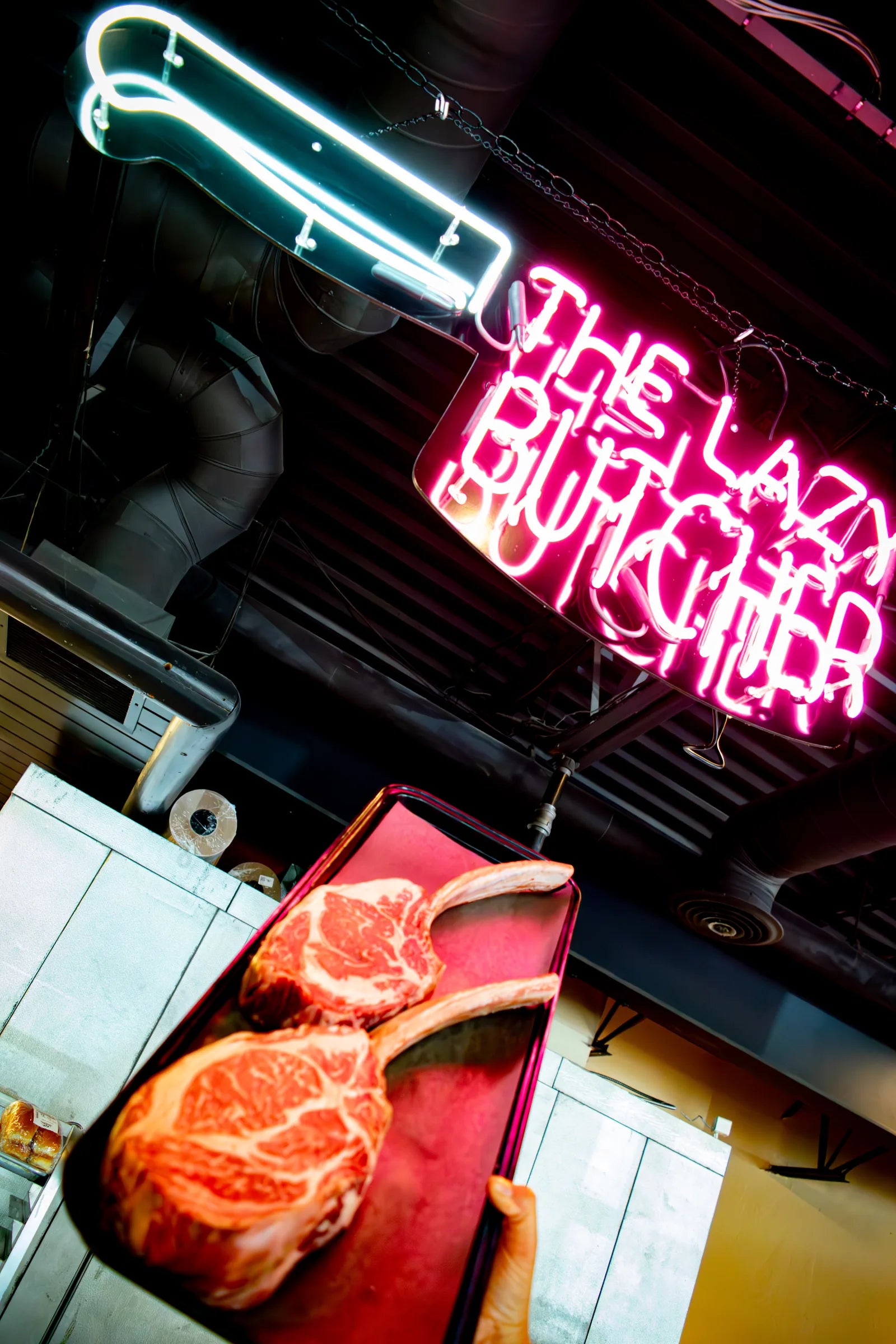 Low-angle shot of two raw bone-in tomahawk steaks with heavy marbling on a red tray, held up beneath a glowing pink and white neon sign reading 'The Lazy Butcher'.