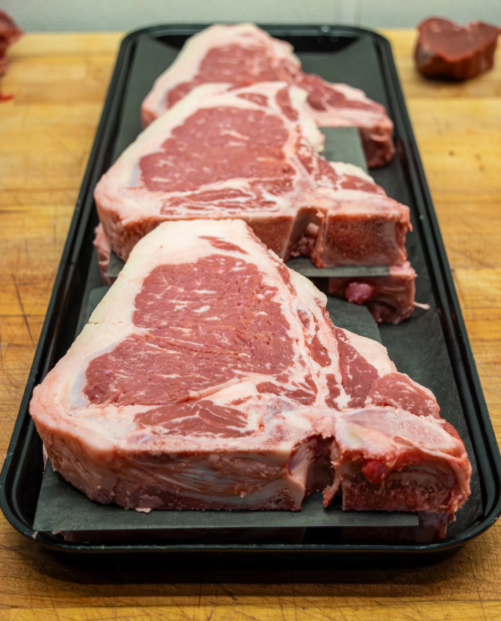 Three thick, heavily marbled porterhouse steaks arranged in a row on a black tray on a wooden butcher block.
