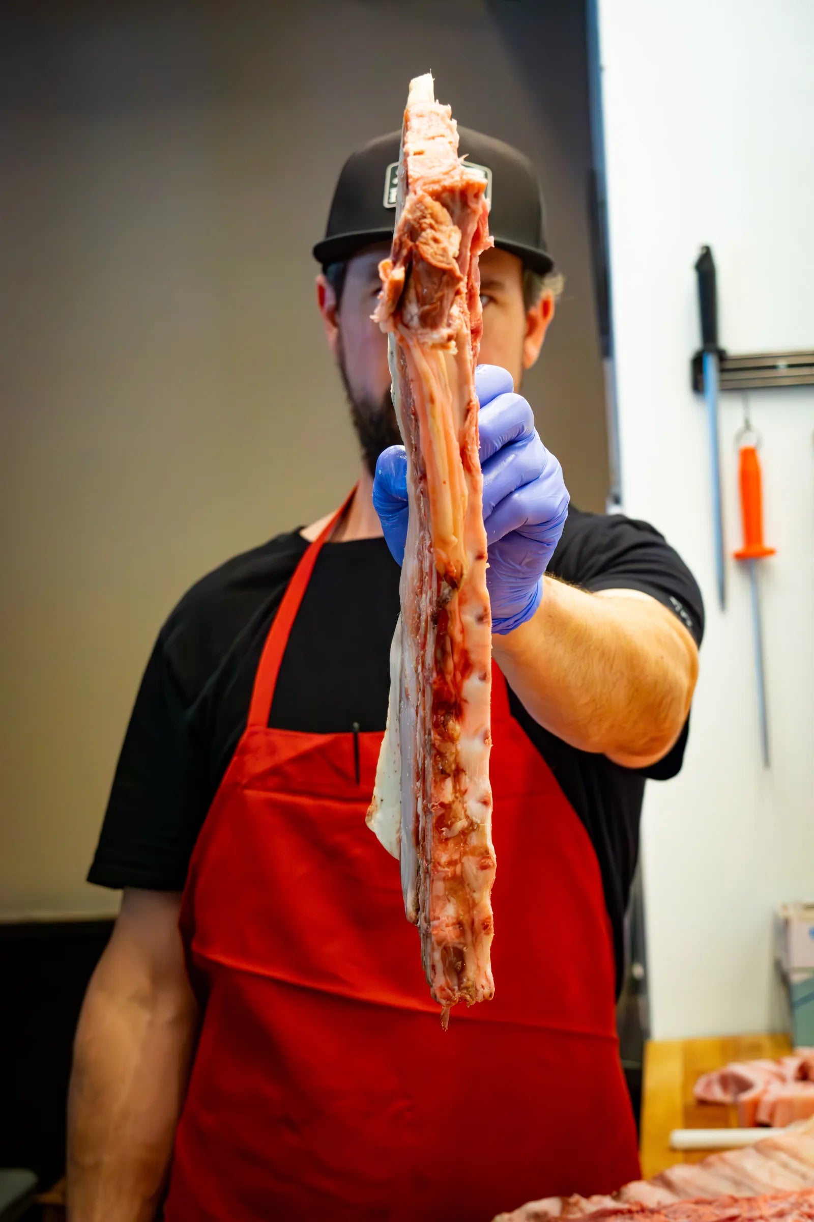 Butcher in black cap and red apron holding up a long trimmed rib bone strip, with knives and meat on the cutting table behind him.