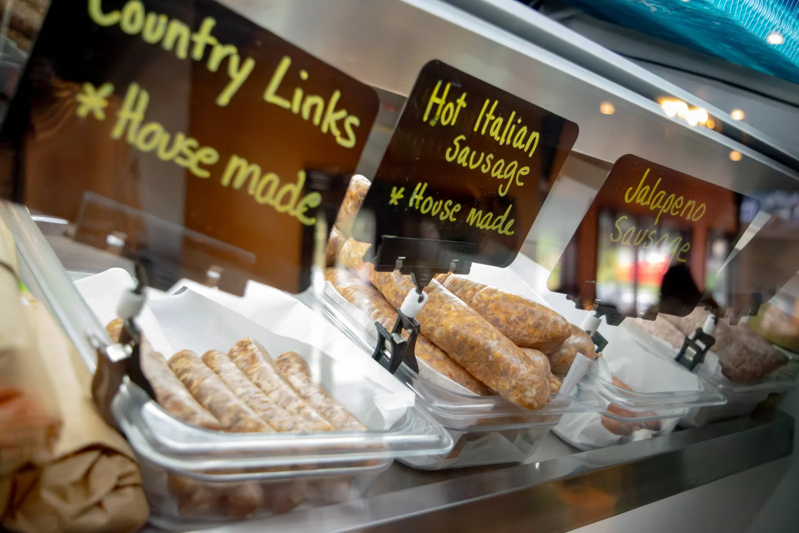 Butcher case display with hand-lettered signs for country links, hot Italian sausage, and jalapeno sausage, with coiled and linked raw sausages beneath each sign.