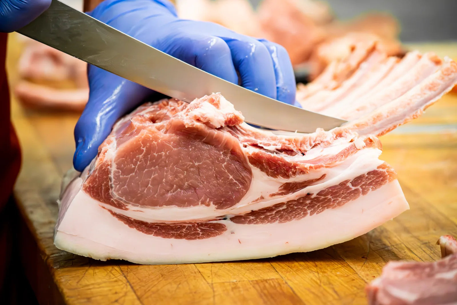 Extreme close-up of a gloved hand using a knife to trim the fat cap from a marbled raw pork chop.