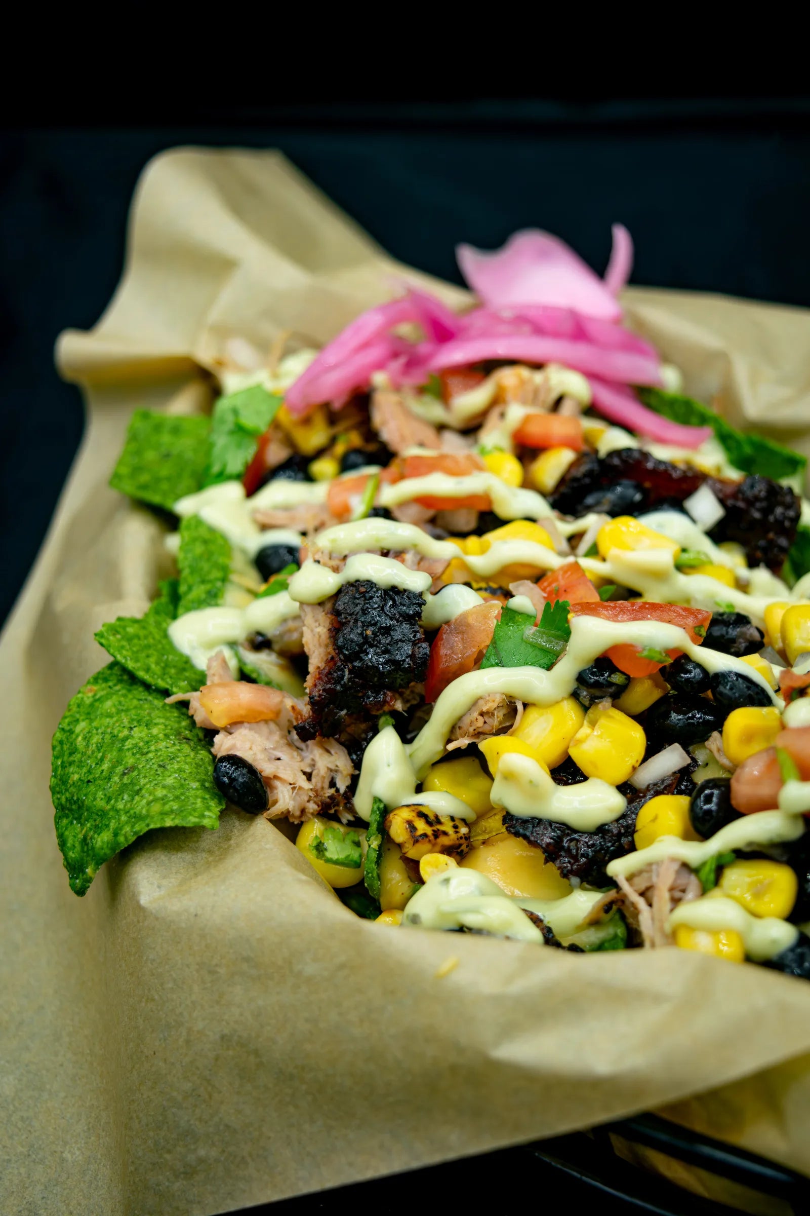 Close-up of nachos made with green tortilla chips, topped with pulled pork, chopped burnt ends, black beans, corn, and avocado crema.