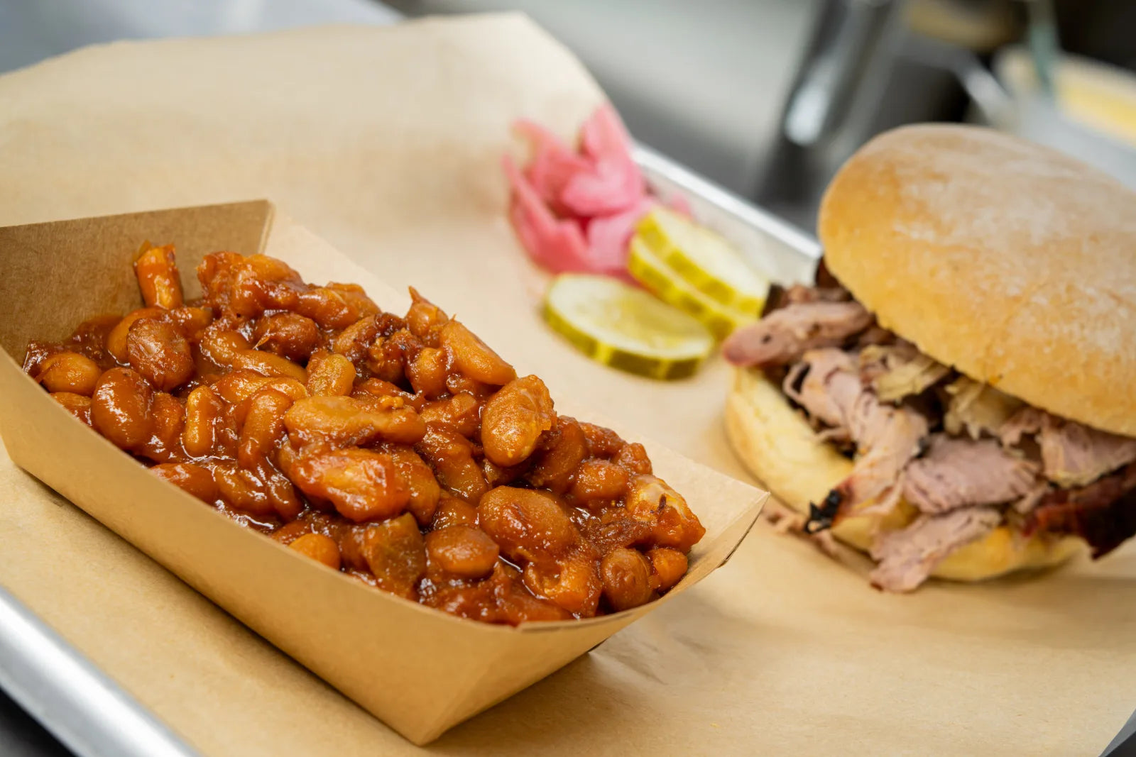 Toasted bun piled with sliced smoked meat next to a paper tray of baked beans and pickle slices.
