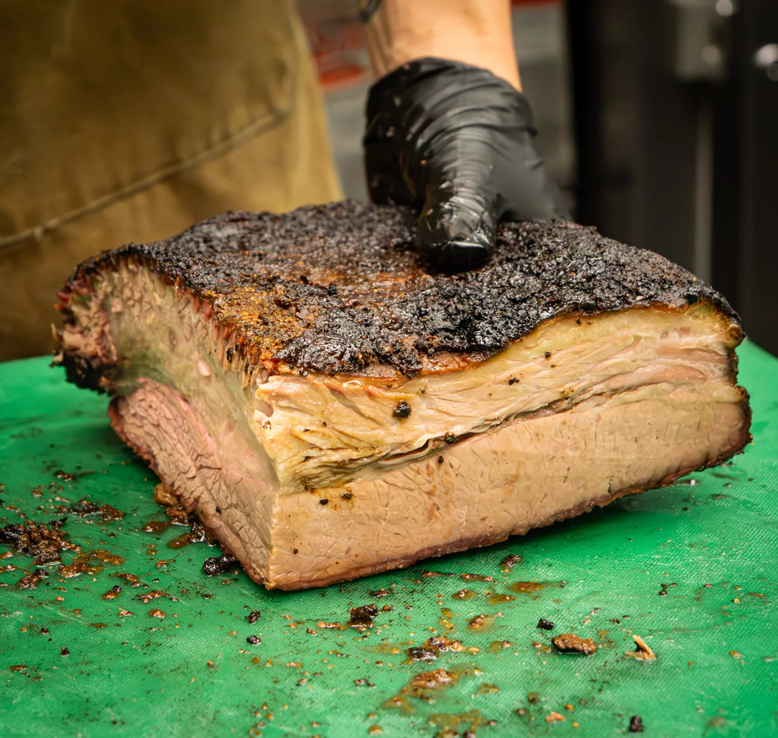 Gloved hand holding up a thick square-cut piece of smoked brisket with a dark peppery bark crust.