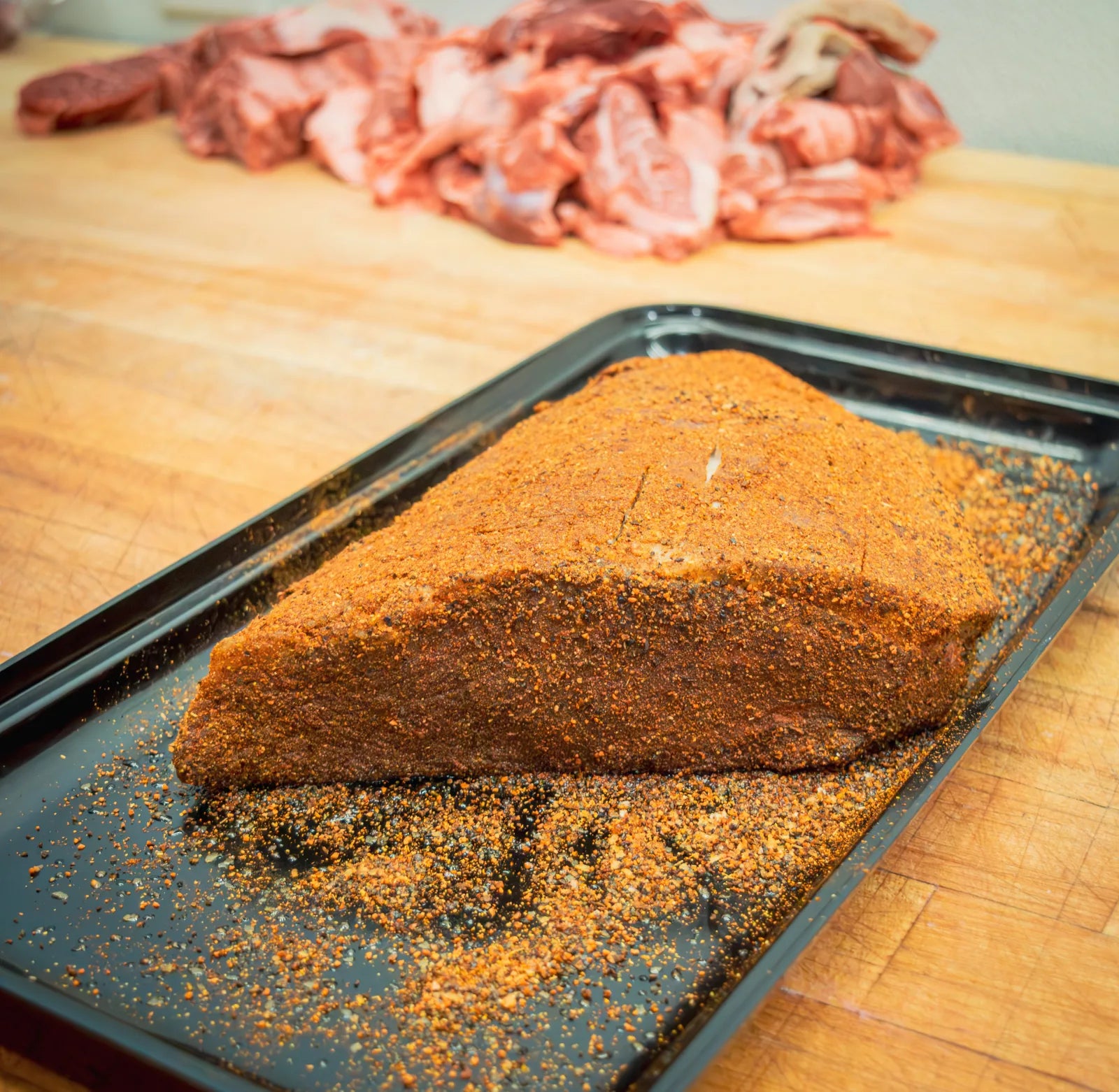 Large cut of beef brisket fully coated in a coarse orange-brown dry rub on a black tray.
