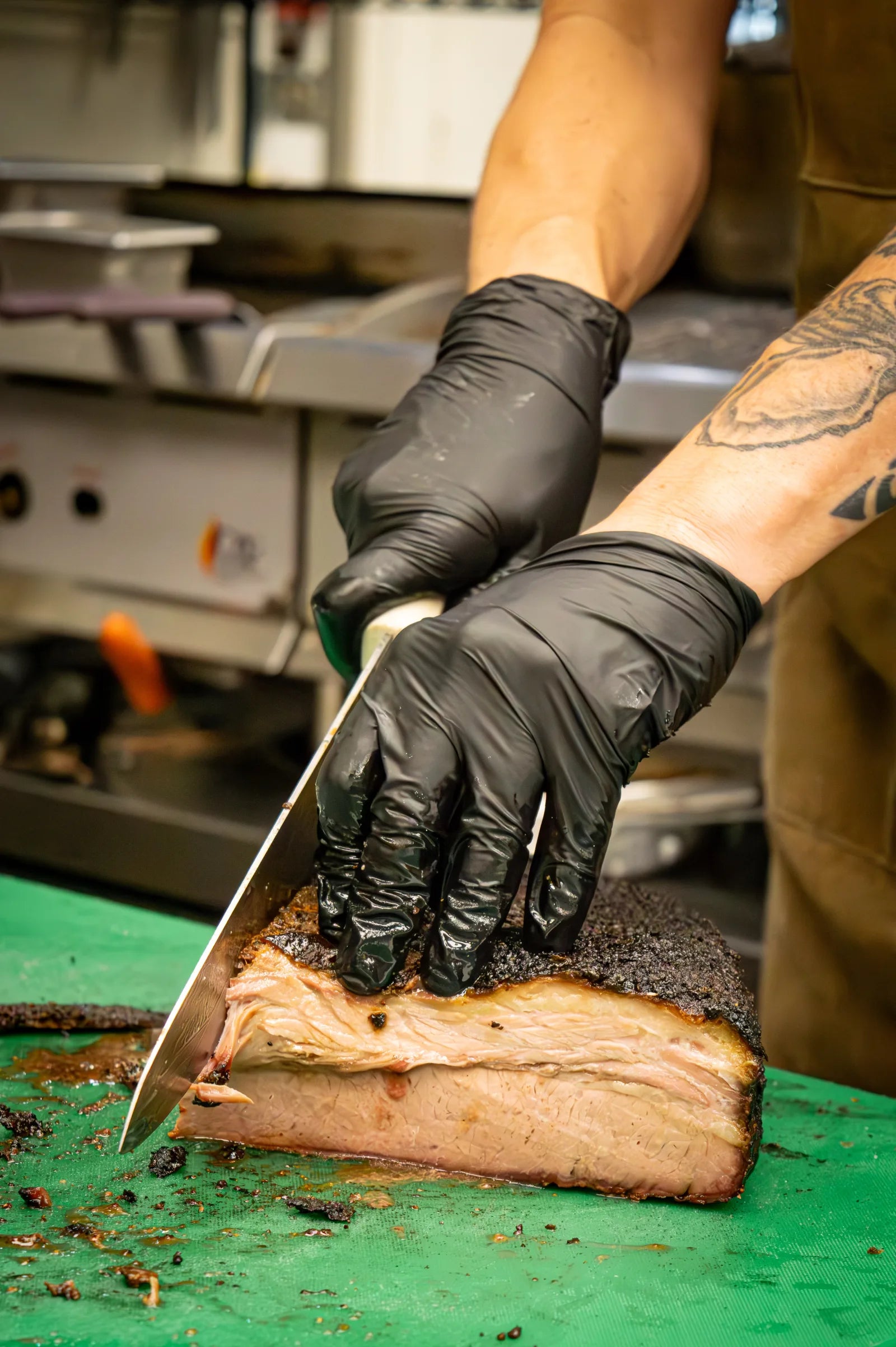 Close-up of gloved hands using a long knife to slice a smoked brisket, showing the dark bark crust and pink smoke ring.