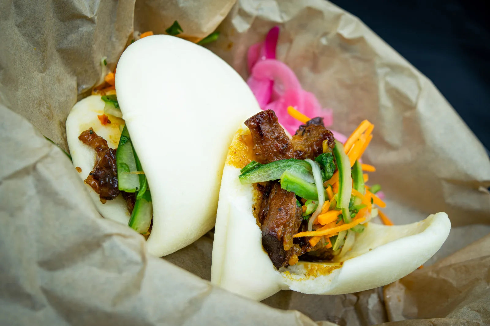 Two open steamed white bao buns filled with glazed burnt ends, shredded carrot, cucumber, and cilantro.