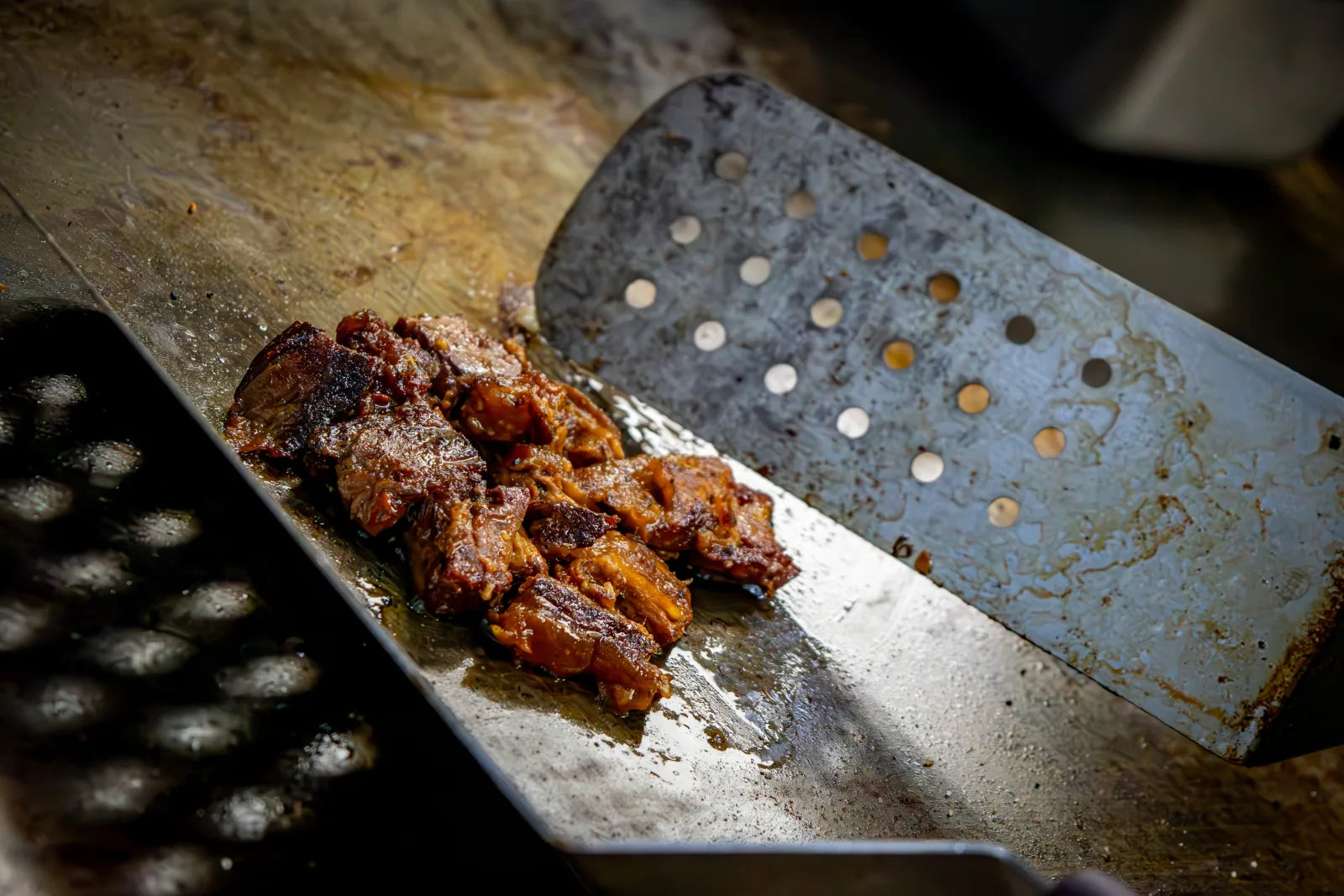 Close-up of caramelized, sauce-glazed burnt ends piled on a perforated metal spatula on a flat top griddle.