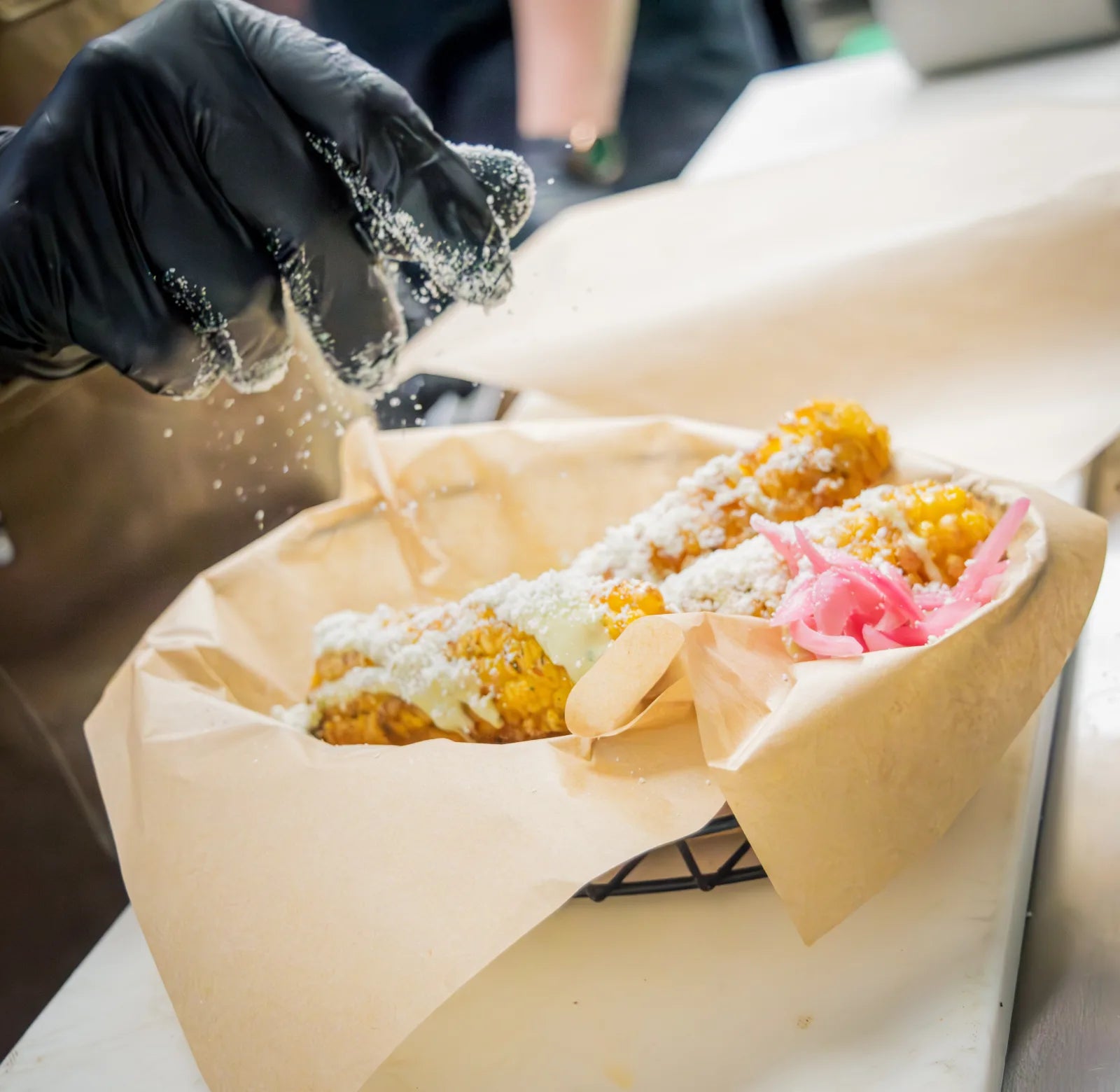 Gloved hand sprinkling crumbled cotija cheese over grilled corn ribs topped with crema and pickled red onion.