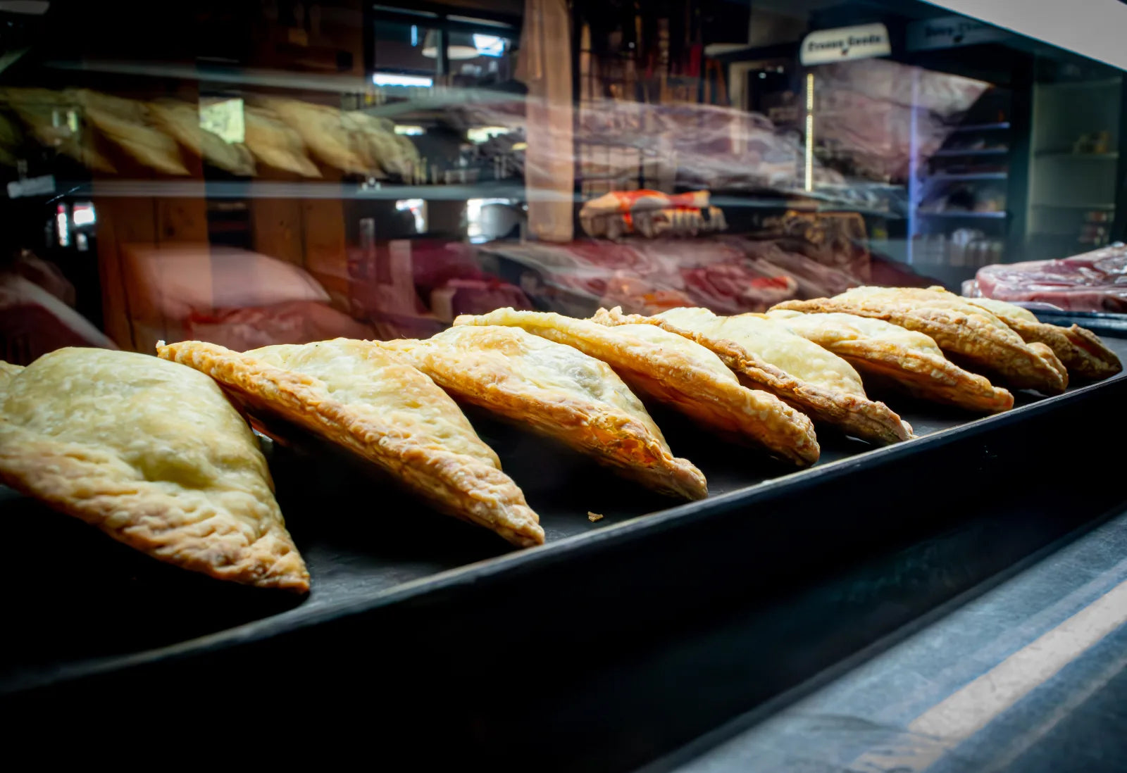 Row of golden-brown baked empanadas lined up in a black display tray, with the meat case reflected in the glass above.