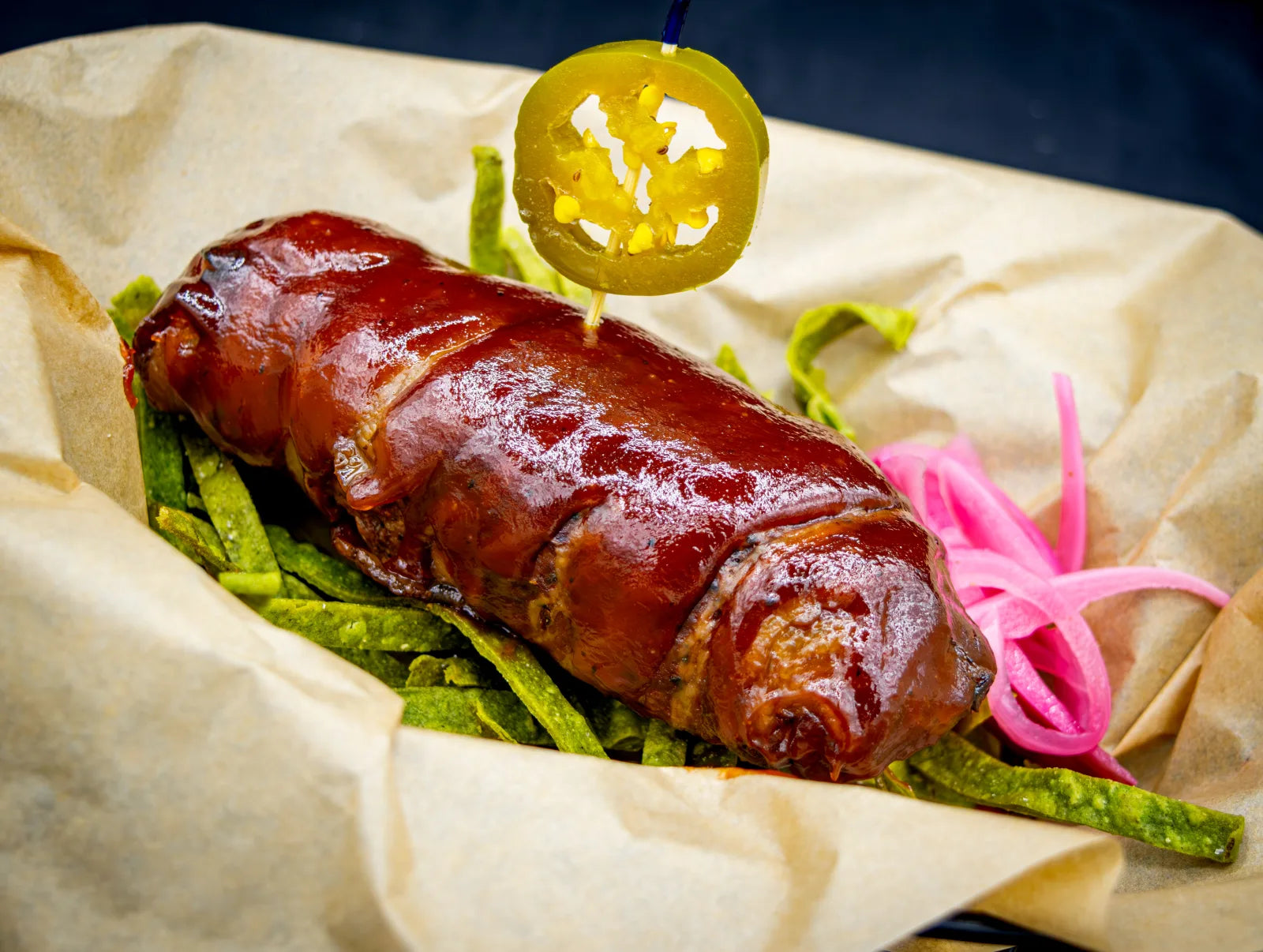 Close-up of a glazed, bacon-wrapped sausage roll with a candied jalapeno slice on a pick, resting on green tortilla strips.