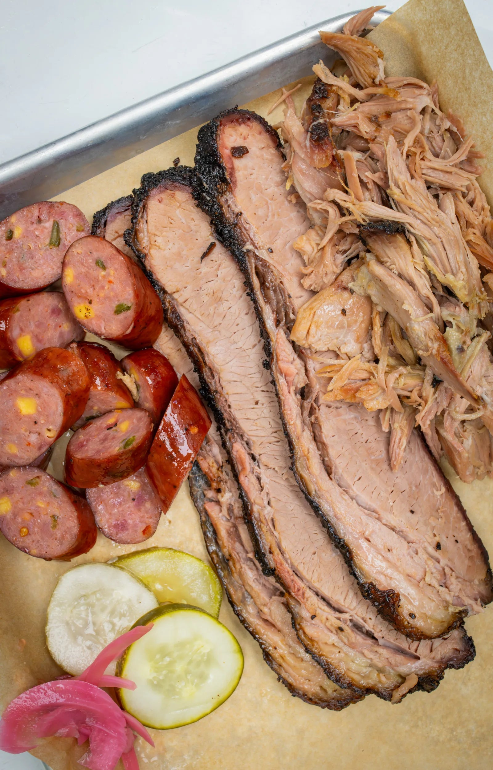 Overhead flat-lay of sliced smoked sausage rounds, sliced brisket, and pulled pork arranged on parchment paper with pickles.
