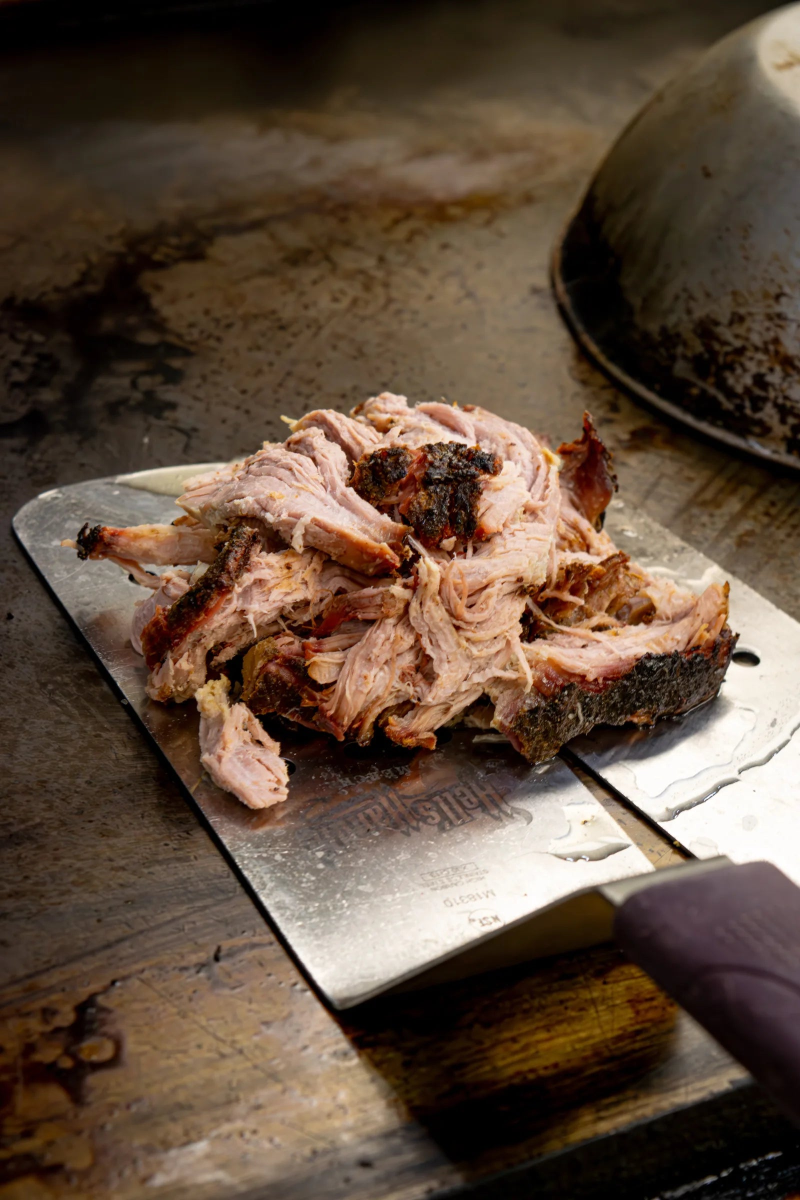 Pile of shredded, bark-flecked pulled pork resting on a large stainless cleaver blade on a scarred wooden butcher block.