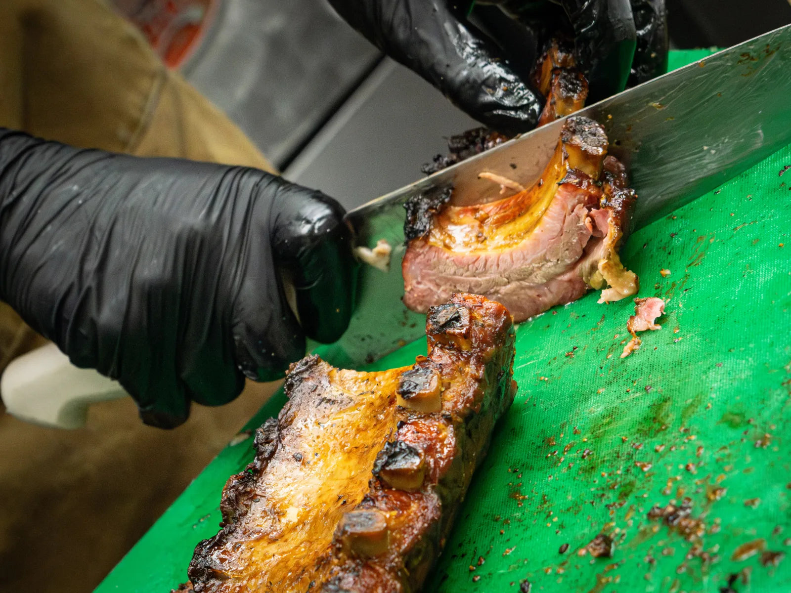 Close-up of gloved hands using a large knife to portion a glazed rack of smoked ribs on a green cutting board.