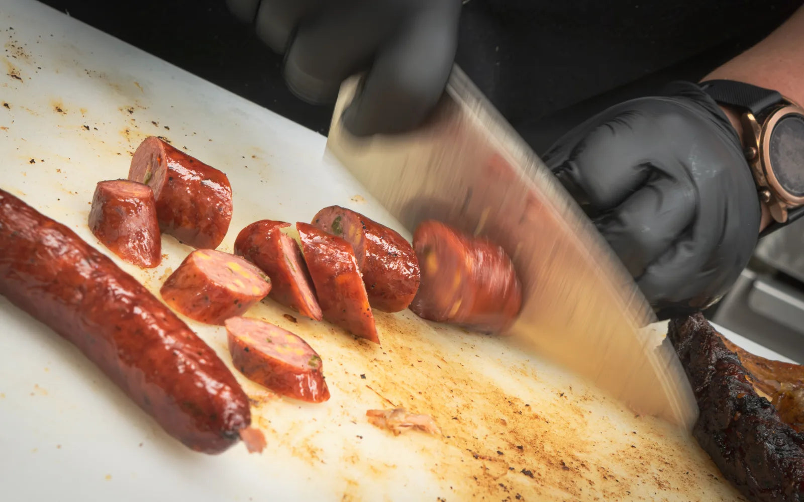 Gloved hand slicing a glazed smoked sausage link into rounds with a chef's knife on a white cutting board.