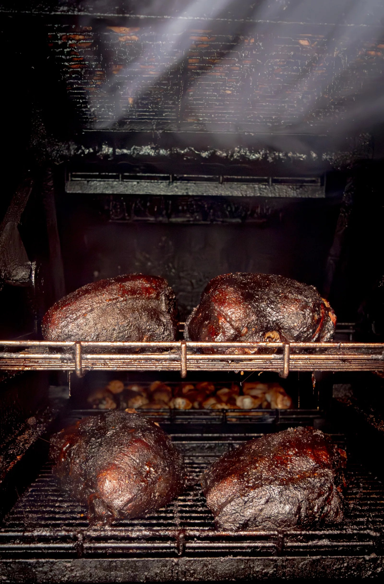 Four whole smoked briskets with dark bark on two racks inside a smoker, with chicken wings smoking below and beams of light cutting through drifting smoke.