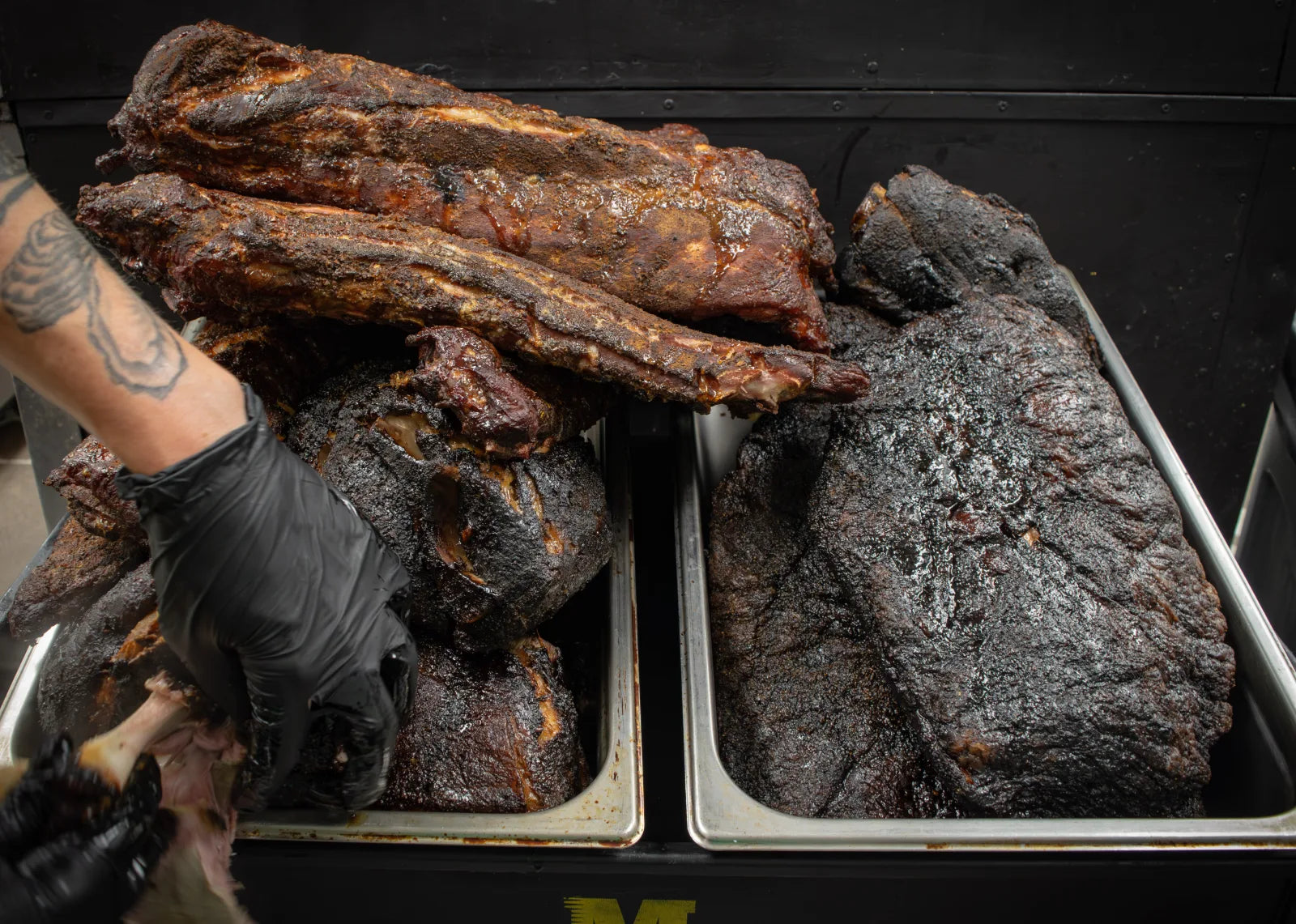 Overhead view of two full metal hotel pans loaded with dark bark-crusted smoked rib racks and whole briskets.