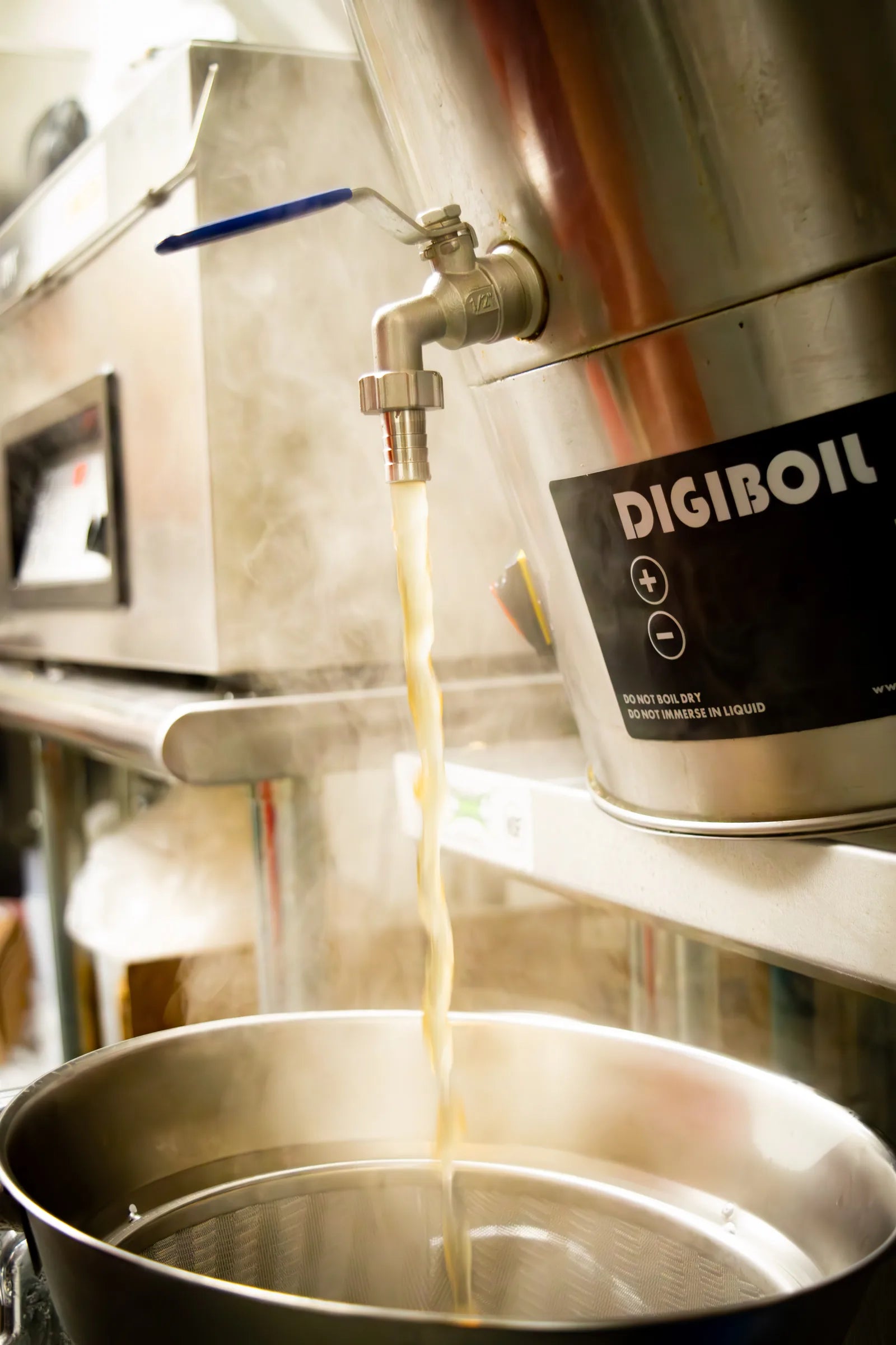 Steaming amber stock pouring from a stainless kettle spigot through a fine mesh strainer, with steam rising in the back kitchen.