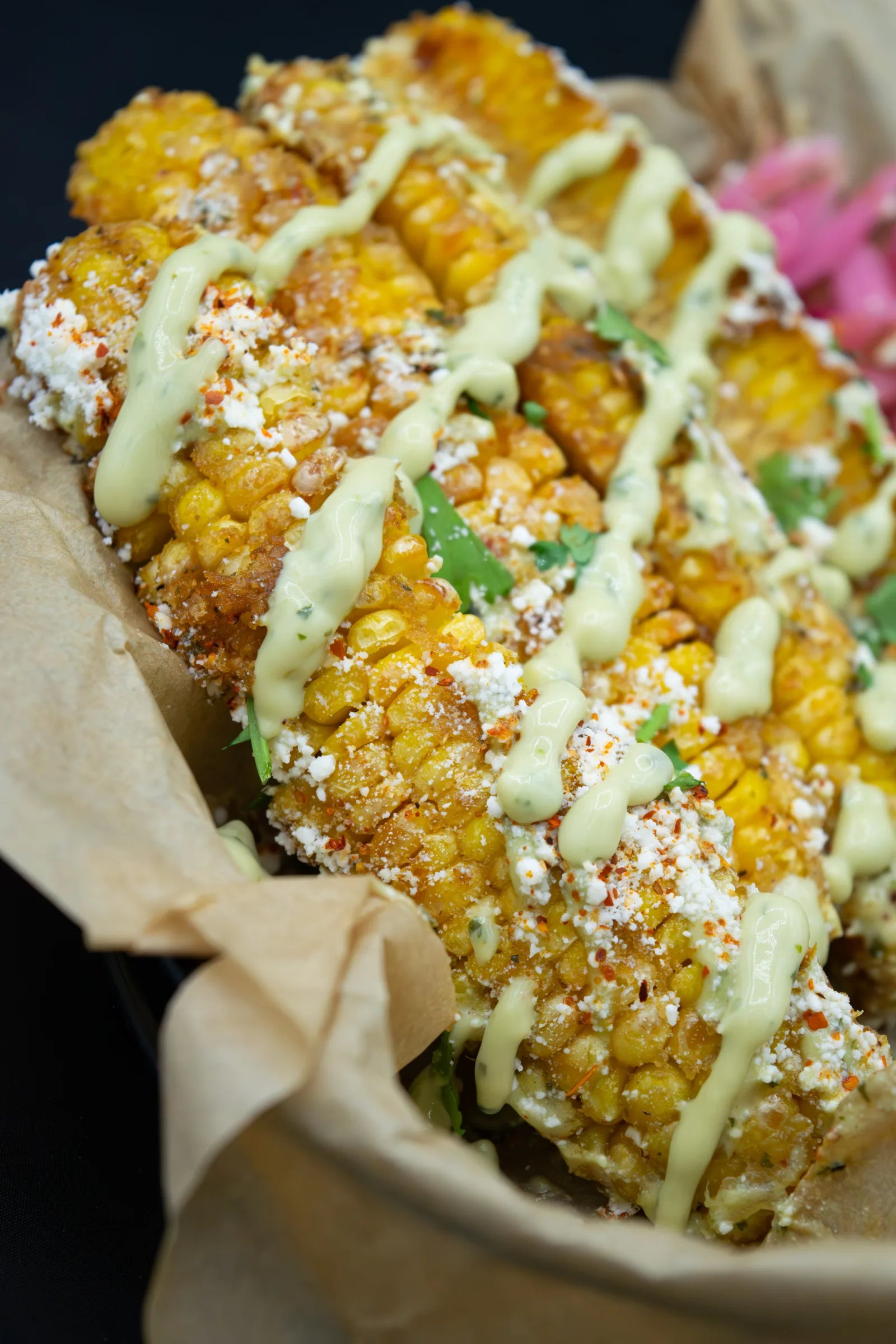Close-up of grilled corn cut into strips, drizzled with pale green crema and topped with crumbled cotija cheese, red chili powder, and pickled red onion.