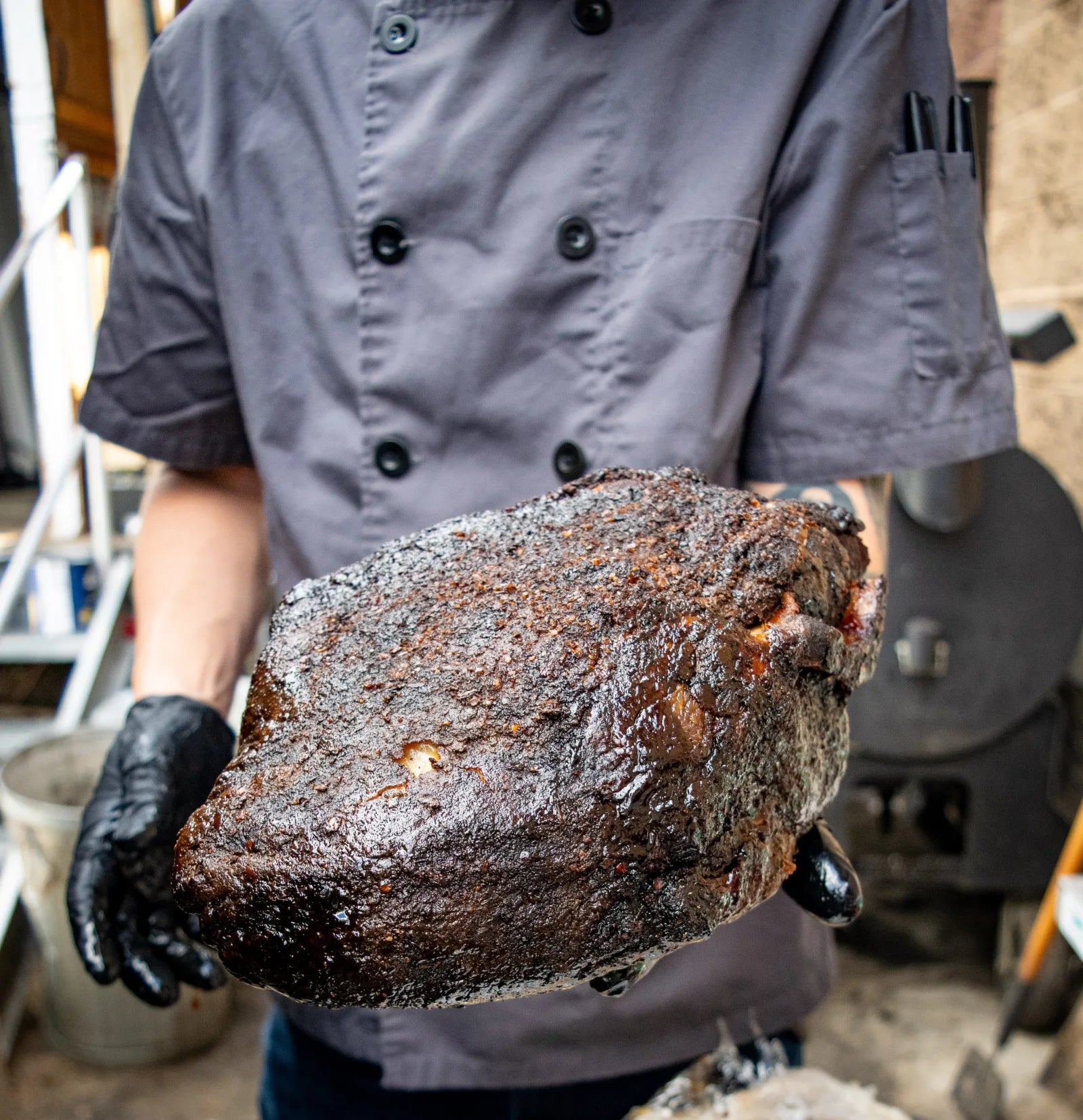 Cook in a gray chef coat and black glove holding out a whole smoked brisket with a dark, glossy bark crust, standing outdoors near a smoker.
