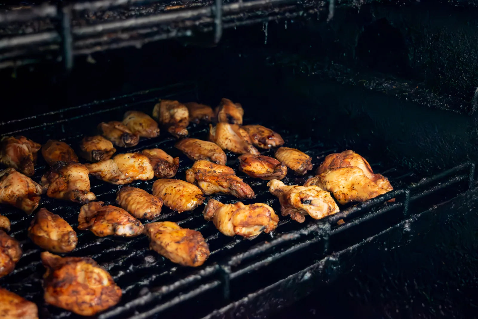 Rows of chicken wings and drumettes cooking on dark smoker grates, lit from the side inside the smoker chamber.