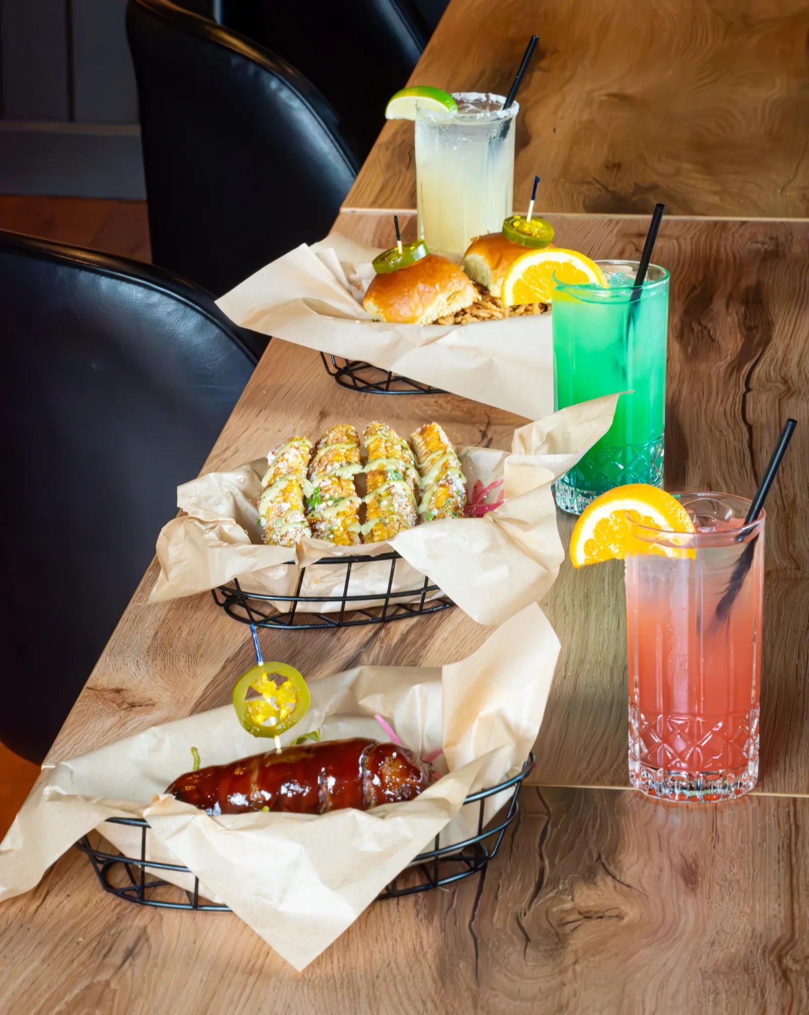 Live-edge wood table set with a basket of BBQ sliders, a basket of grilled corn, a glazed sausage, and cocktails garnished with citrus.