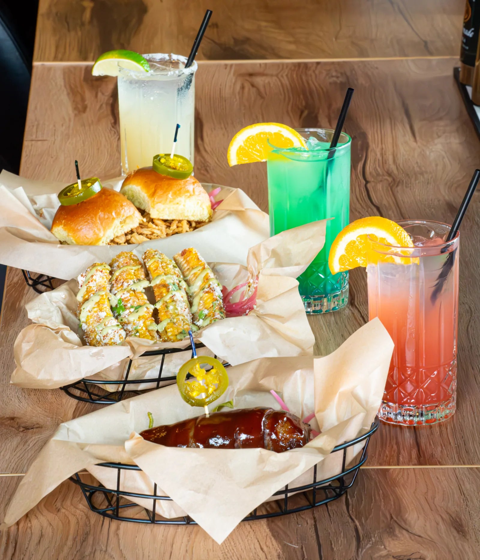 Overhead view of a live-edge wood table set with baskets of BBQ sliders and grilled corn, a glazed sausage, and four colorful cocktails.