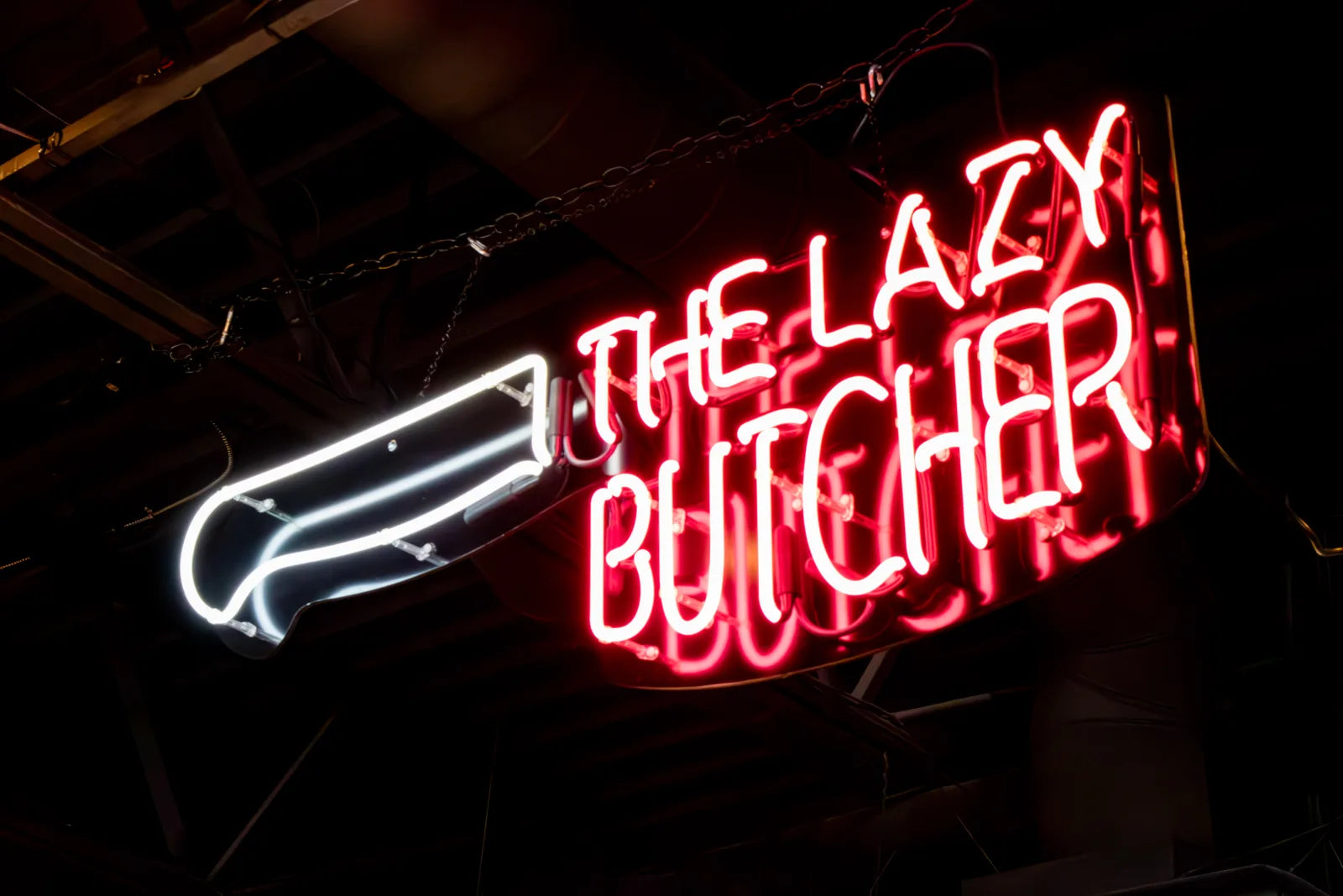 Close-up of a neon sign reading 'The Lazy Butcher' in red script beneath a white neon knife icon, glowing against a black background.