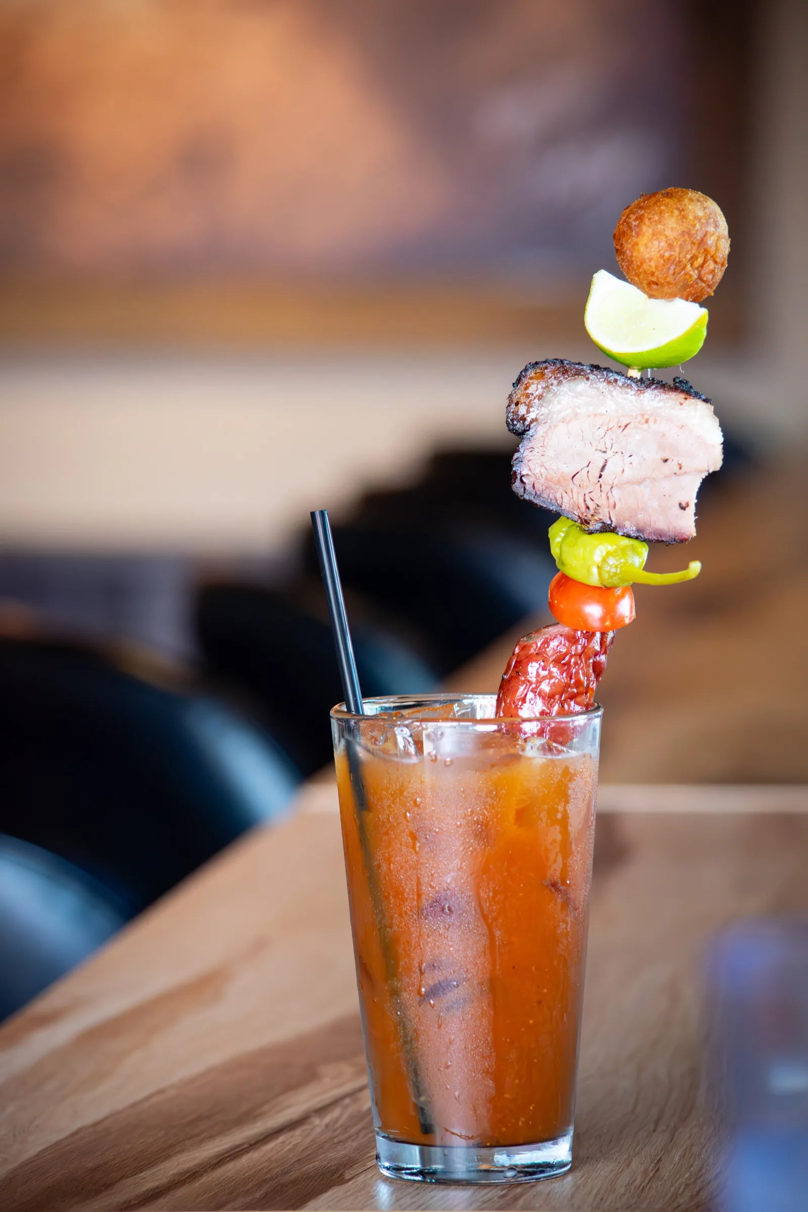 Tall Bloody Mary cocktail with a skewer garnish stacked with a fried hushpuppy, lime wedge, smoked pork belly, a pepperoncini, and a sausage stick.