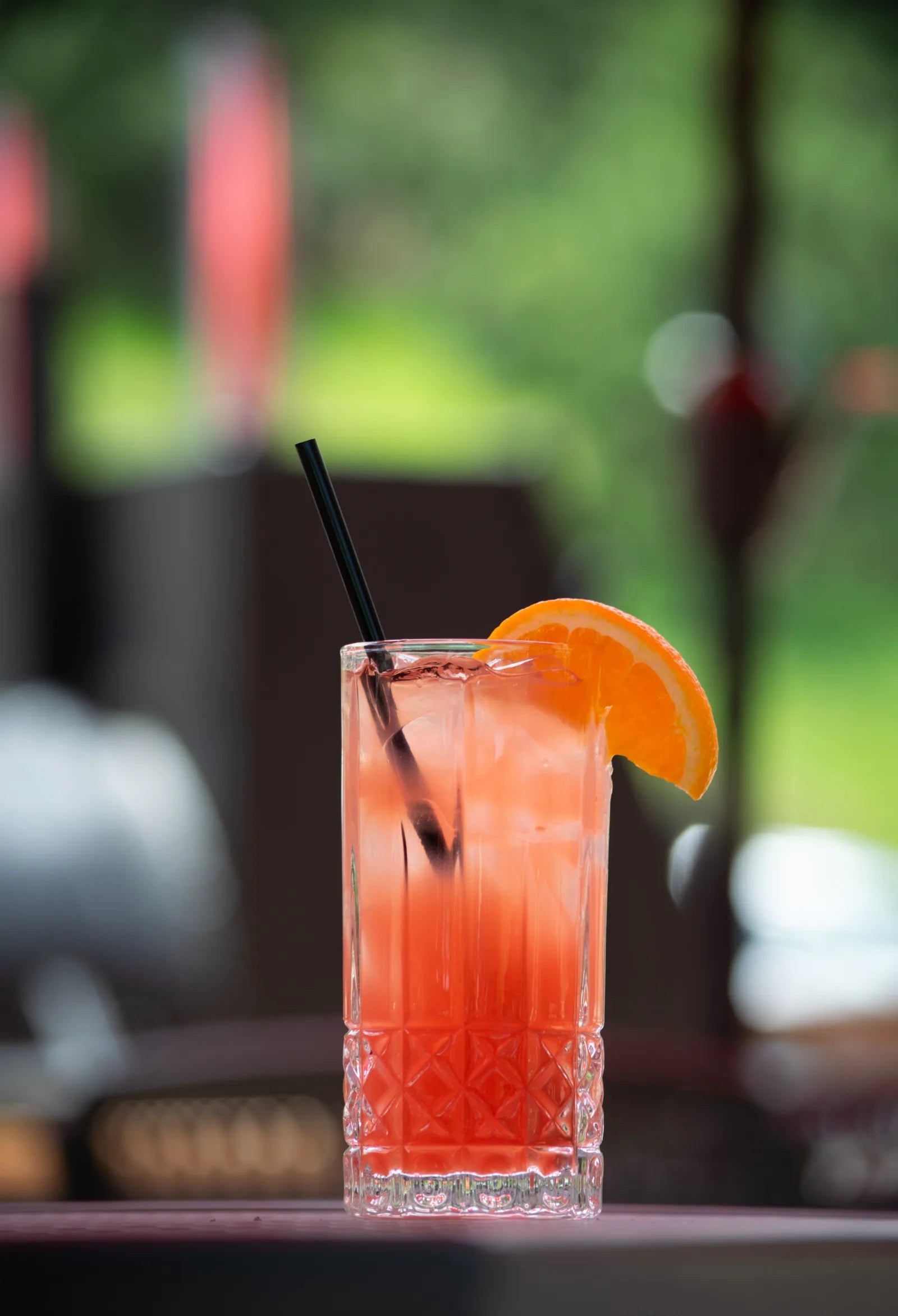 Tall glass with a red-orange cocktail, ice, dark straw, and an orange wheel garnish on the rim.