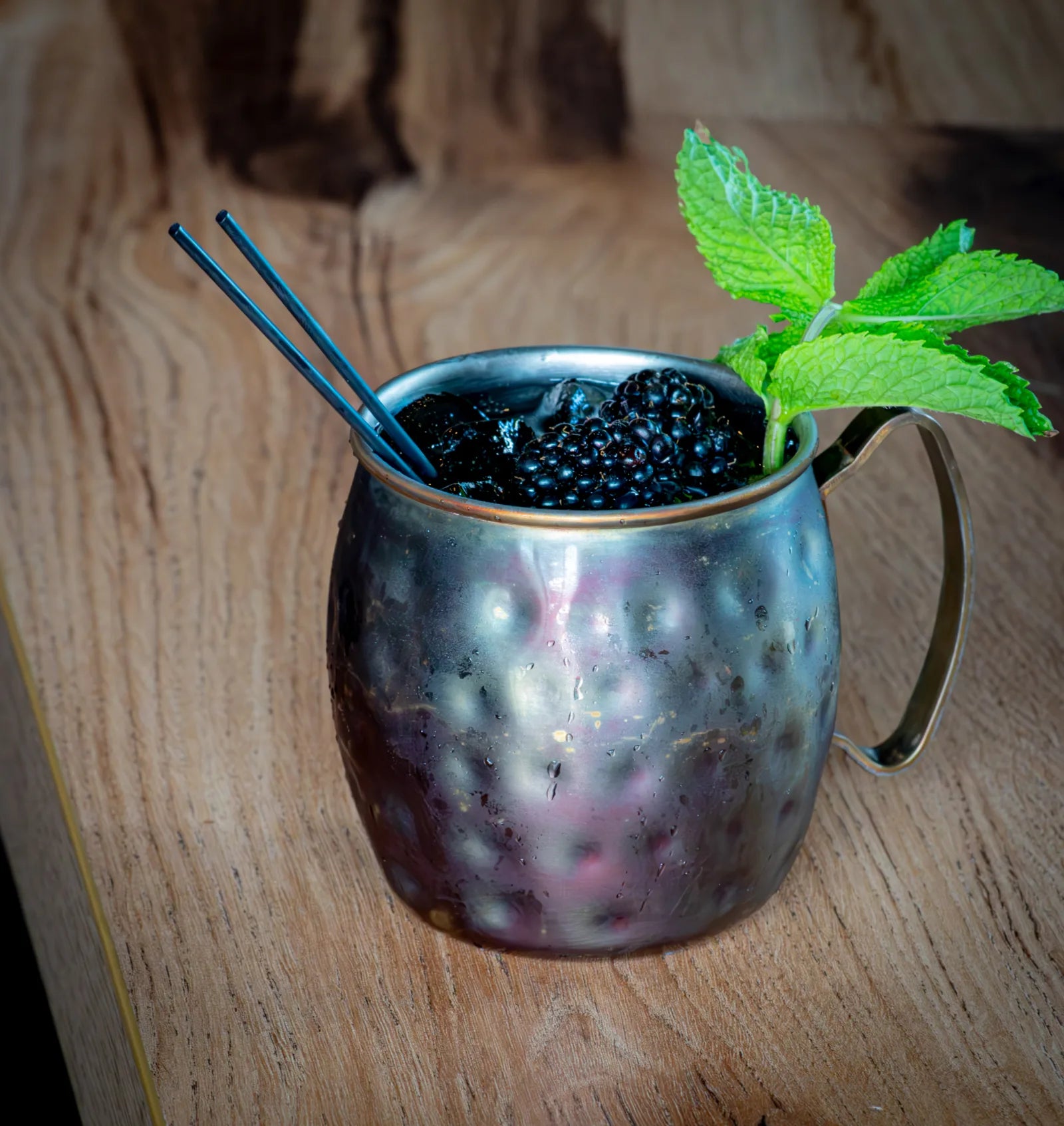 Hammered copper mug filled with a dark cocktail, ice, fresh blackberries, and a mint sprig garnish with two metal straws.