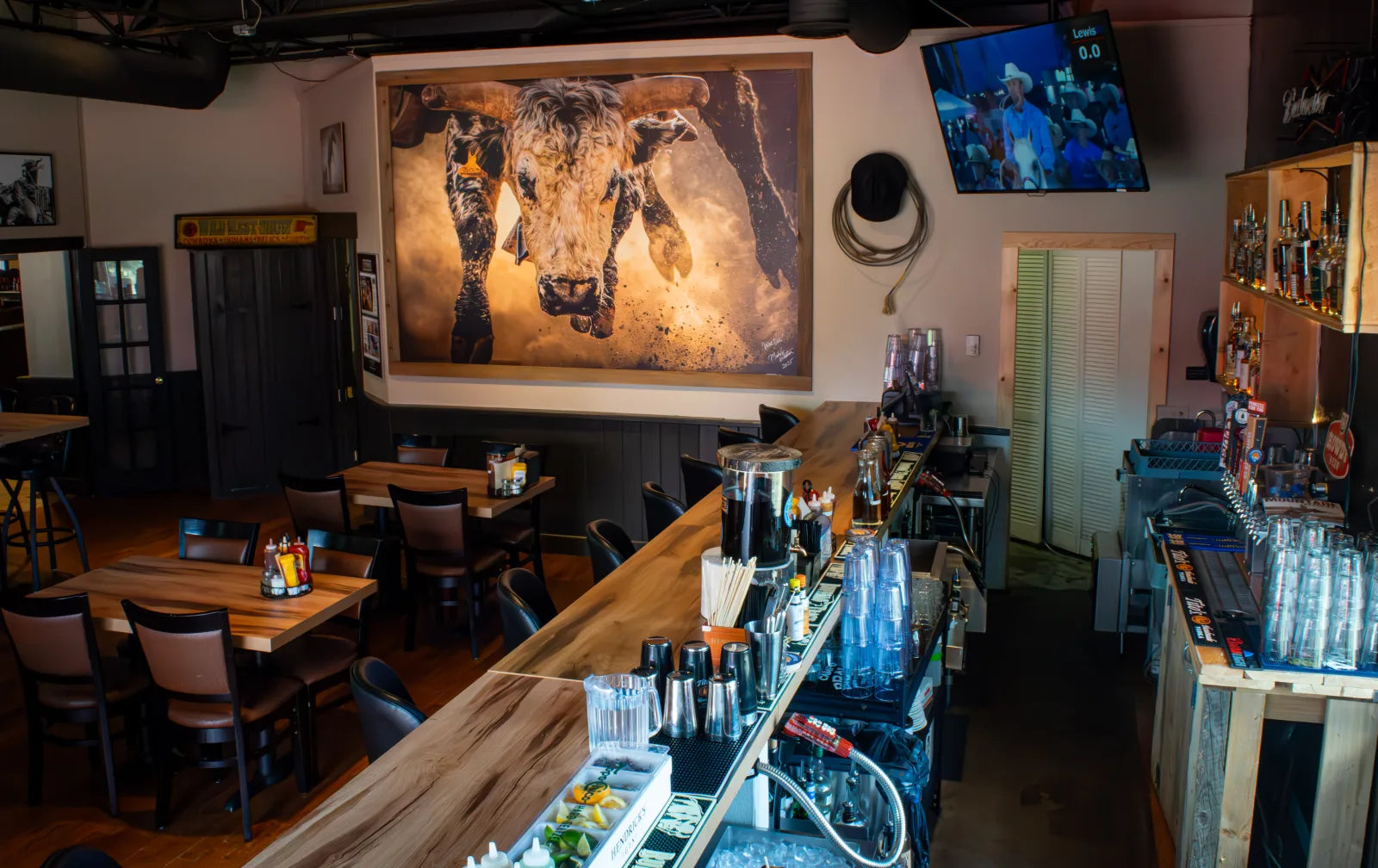 Wide interior view of the Cowboy Lounge showing a live-edge wood bar counter with barstools, dining tables, a large framed photograph of a bucking bull, and a TV playing rodeo.
