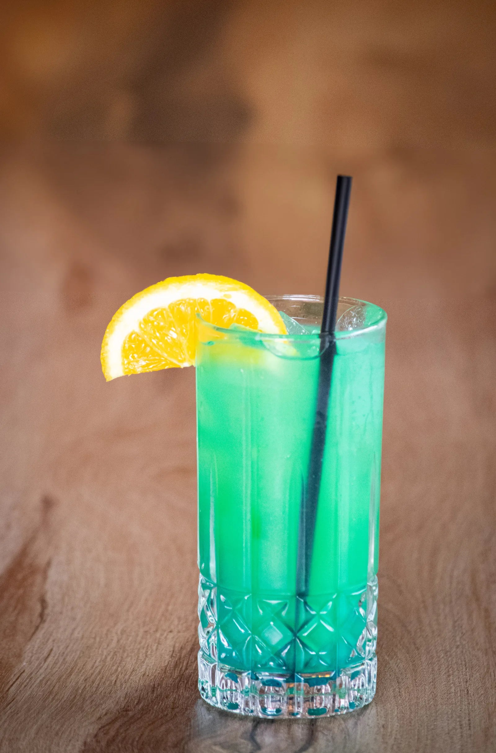 Tall crystal-cut glass filled with a bright green cocktail, ice, black straw, and an orange wheel garnish.