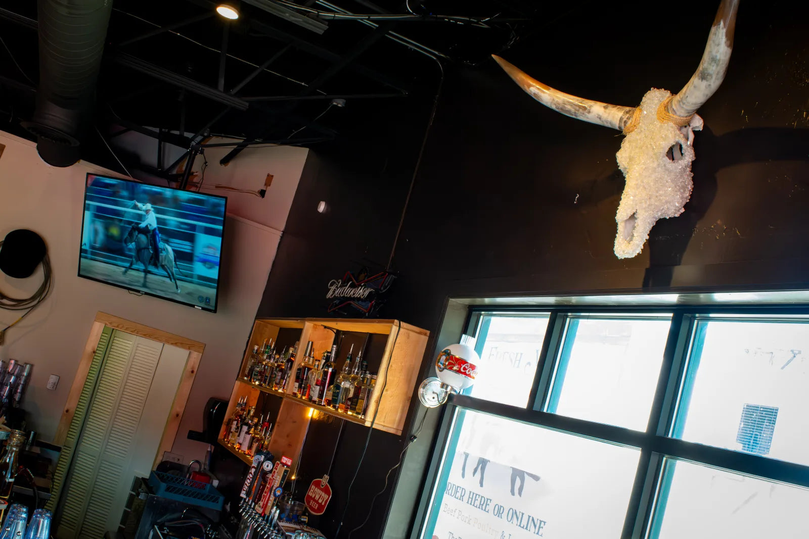 Wide interior shot of the Cowboy Lounge bar looking up at a crystal-covered longhorn skull mounted near the ceiling, with a TV playing rodeo footage.
