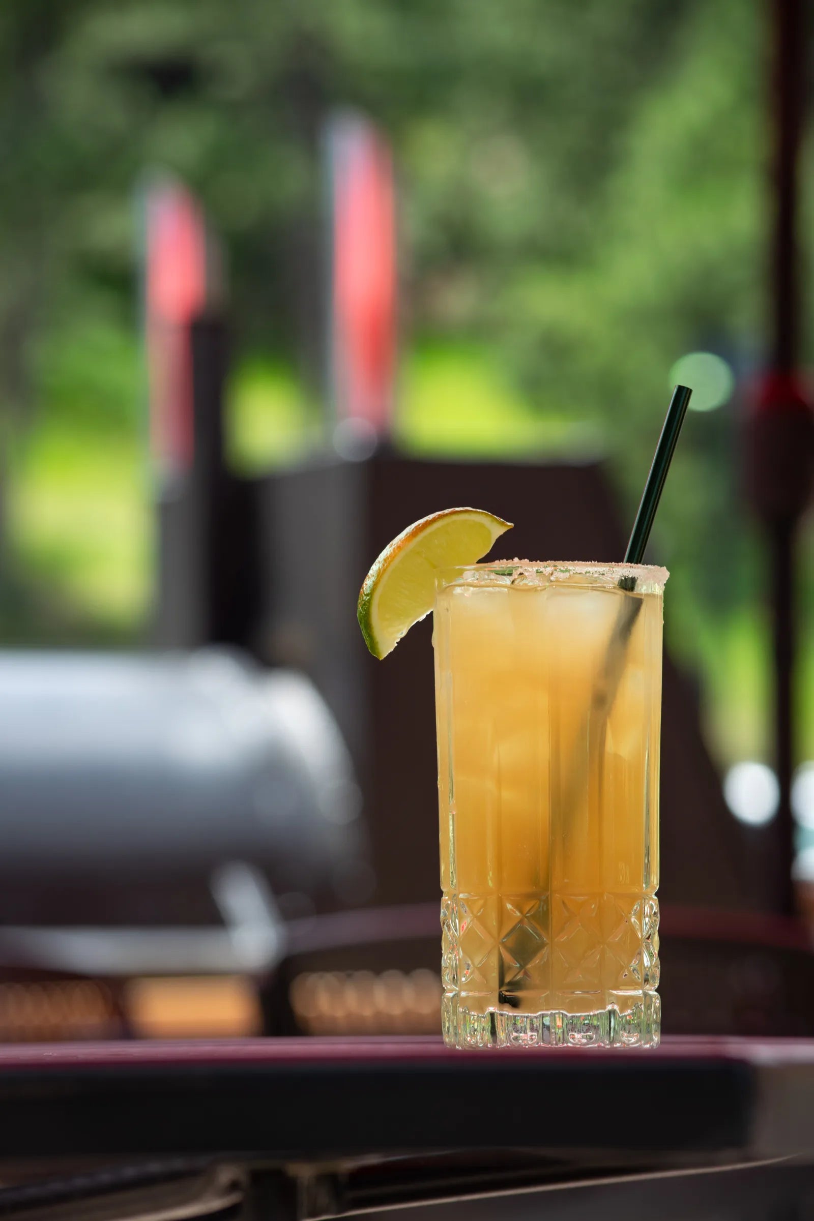 Tall glass cocktail with an orange-hued drink, salted rim, ice, dark straw, and lime wedge garnish, on an outdoor table.