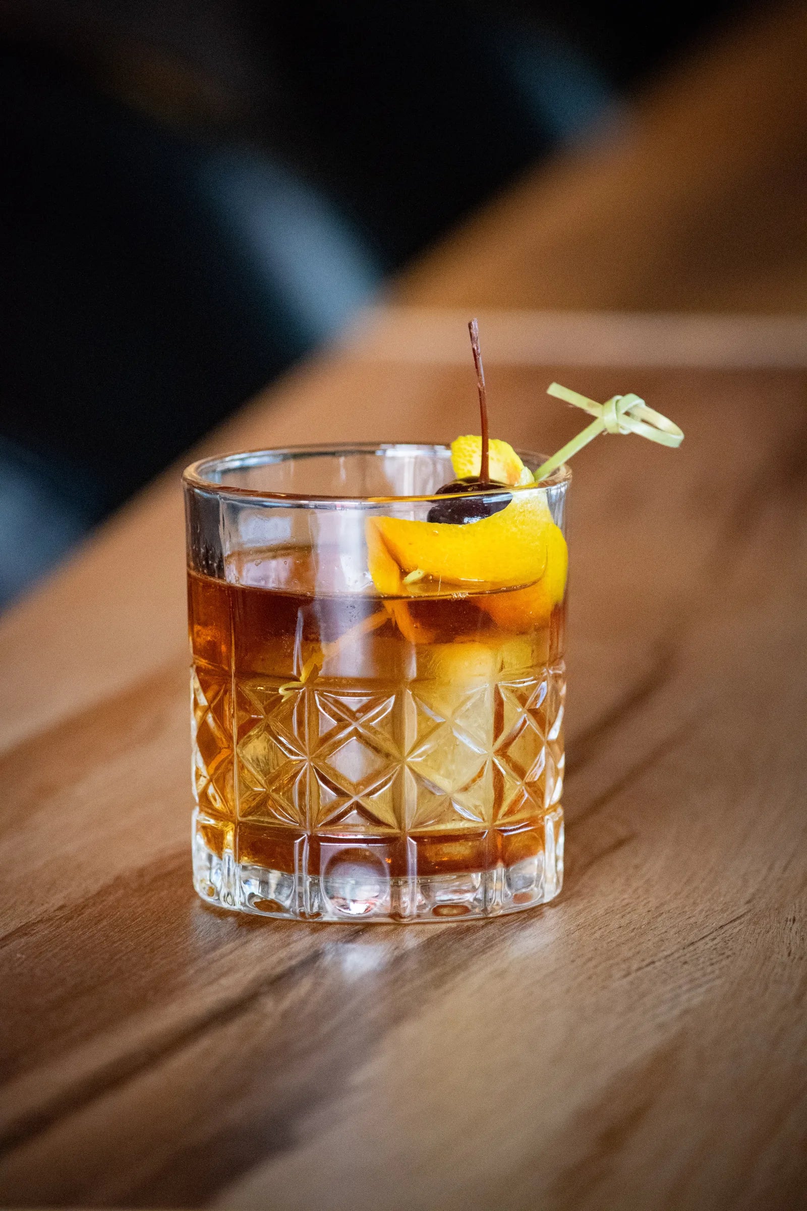 Old fashioned cocktail in a cut crystal rocks glass with a large ice cube, garnished with an orange peel twist and a cocktail cherry.