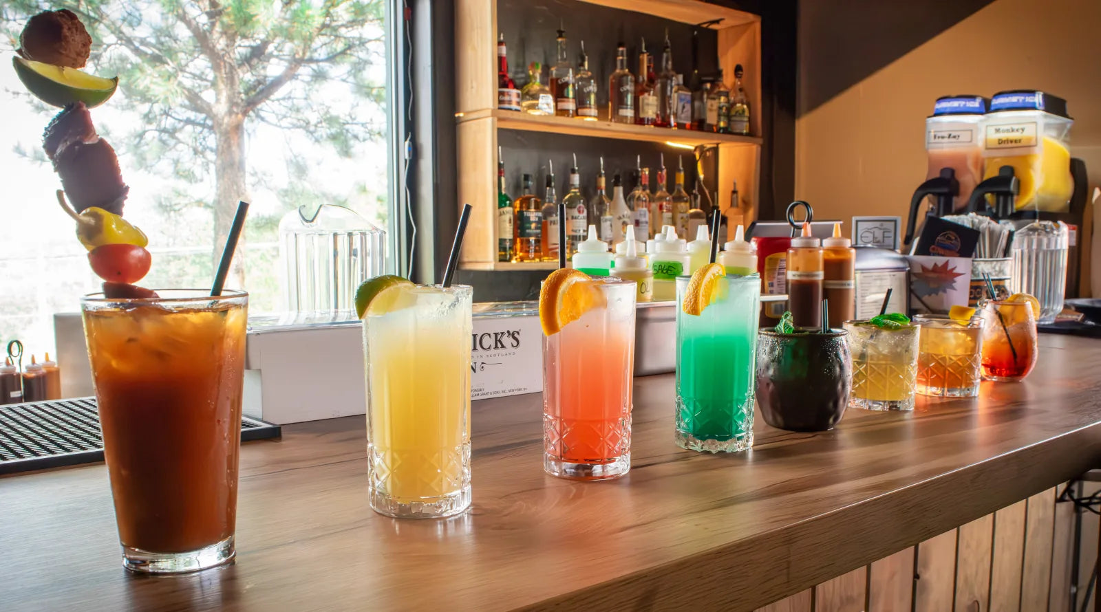 Row of colorful cocktails including a Bloody Mary and margarita lined up on a wood bar counter, with a pine tree and mountain view visible through a large window.