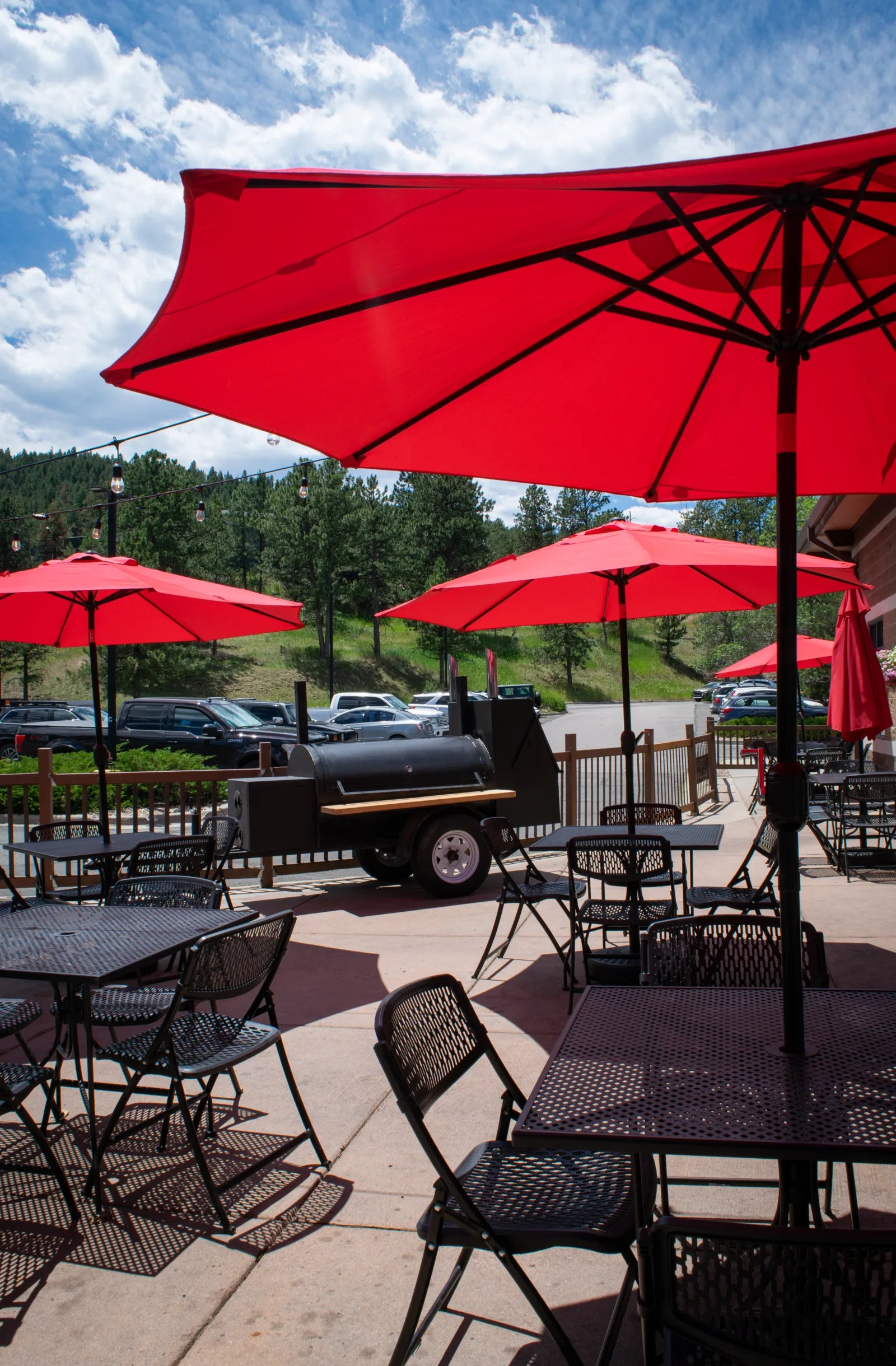 Outdoor patio with black metal tables, chairs, and open red umbrellas; a black barbecue smoker trailer sits behind a wood fence with pine trees and blue sky beyond.
