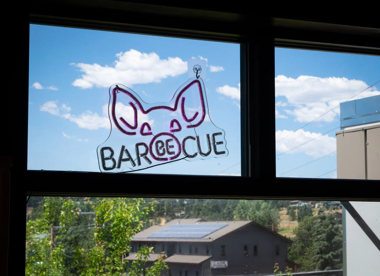 Neon sign shaped like a pig's head reading 'Bar-Be-Cue' mounted in a window, with blue sky and a mountain neighborhood visible through the glass.