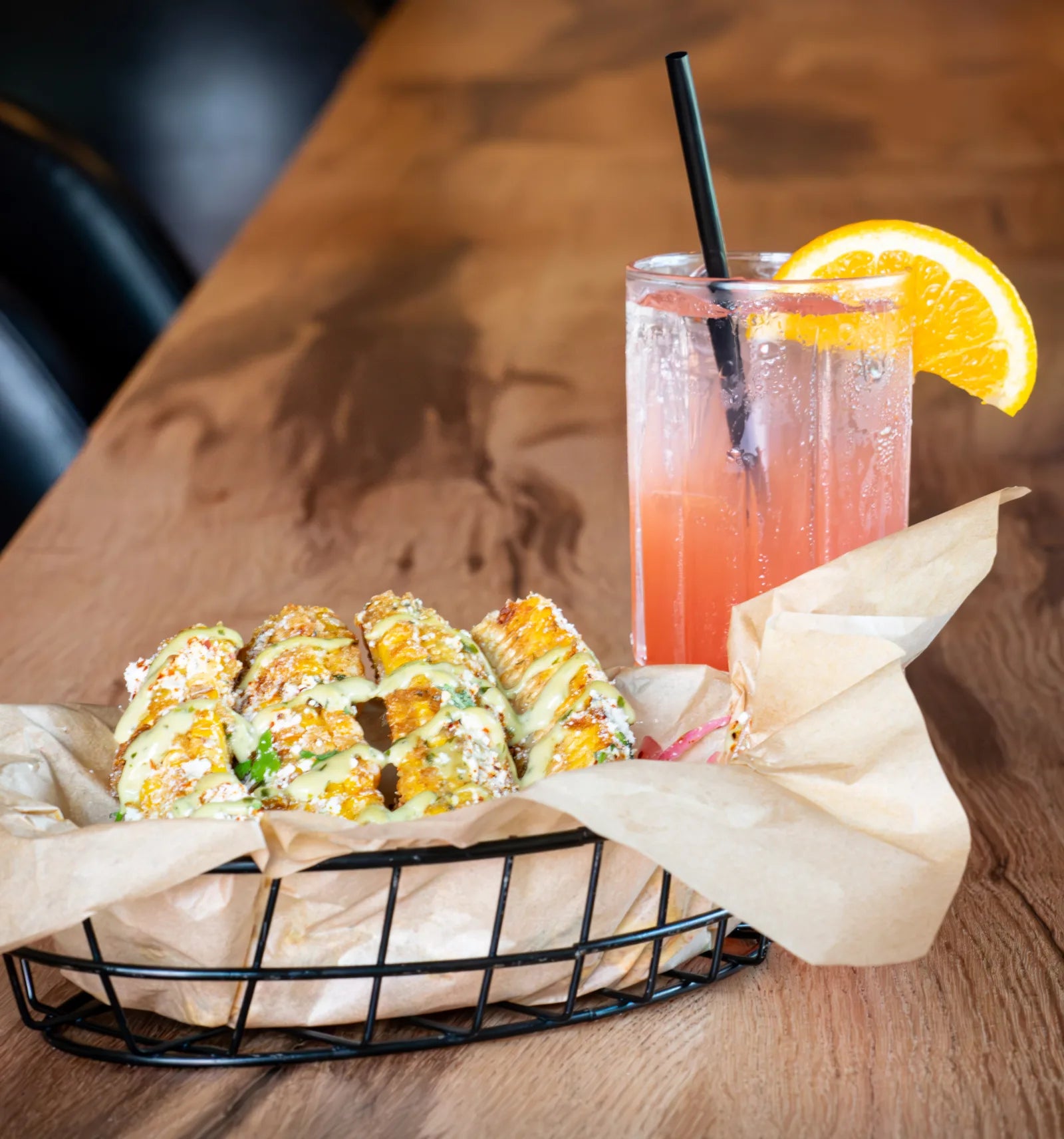 Wire basket of grilled corn topped with crema and cotija cheese next to a tall pink cocktail garnished with an orange wheel.