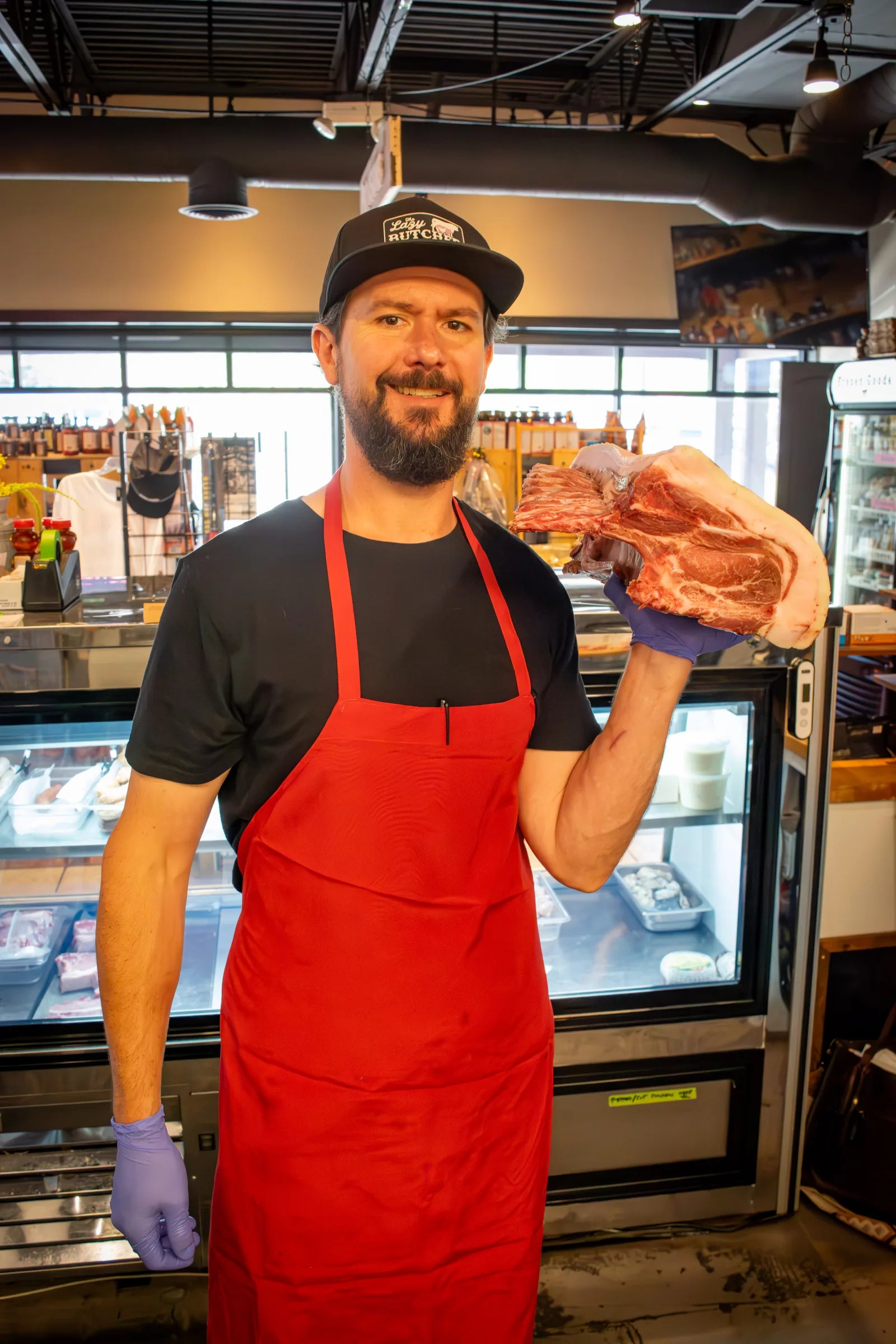 Smiling bearded butcher wearing a black 'The Lazy Butcher' cap and red apron, holding up a large raw rib rack beside the meat case.