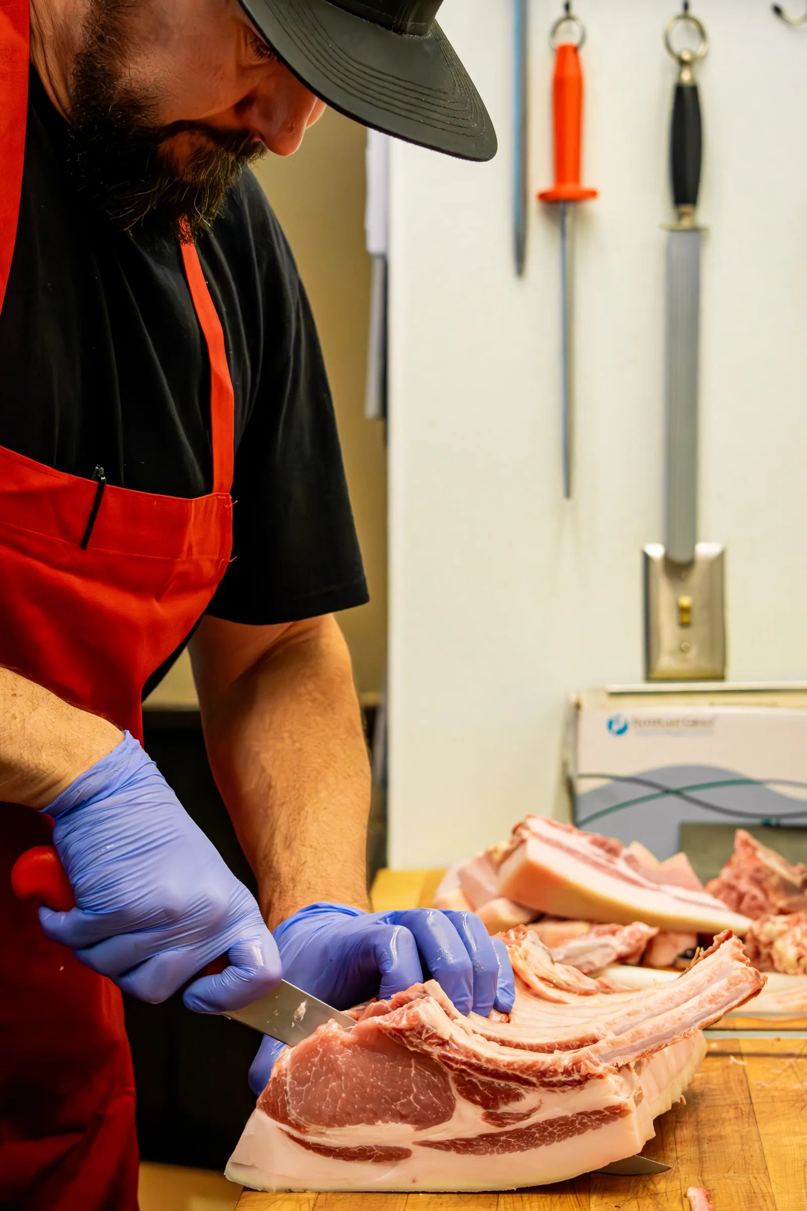 Bearded butcher in a black cap and red apron leaning over a rib rack, with knives and sharpening steels hanging on the wall behind him.