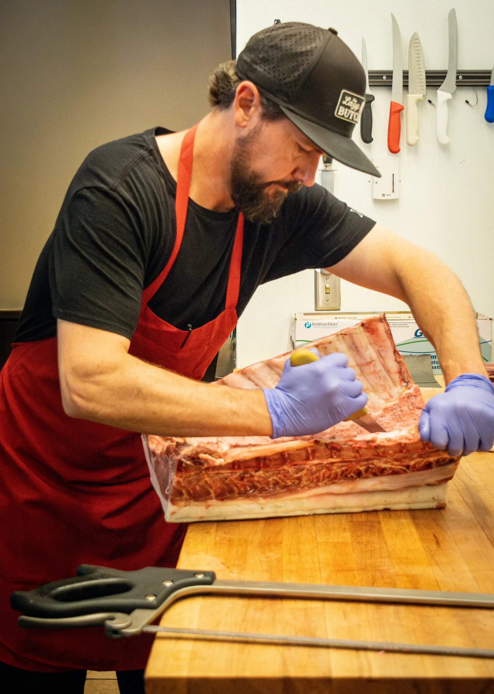 Bearded butcher wearing a black 'The Lazy Butcher' cap and red apron leaning over a workbench trimming a large raw rib rack.