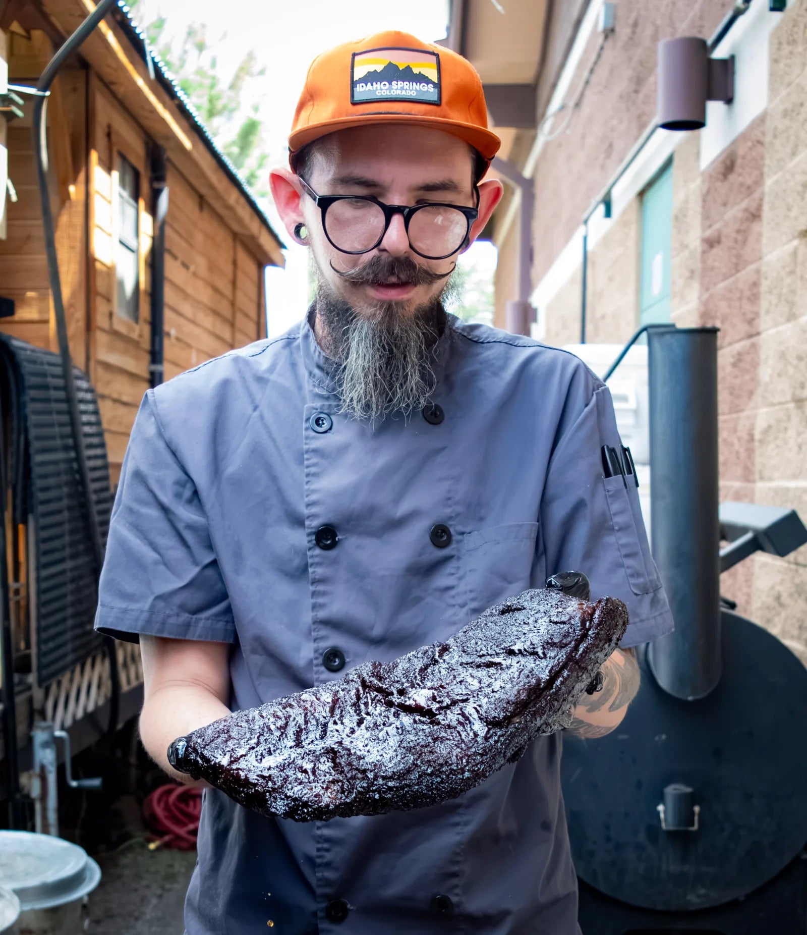 Cook with a long gray beard and orange cap, wearing a gray chef coat, holding out a dark smoked rack of ribs outside near the smoker.