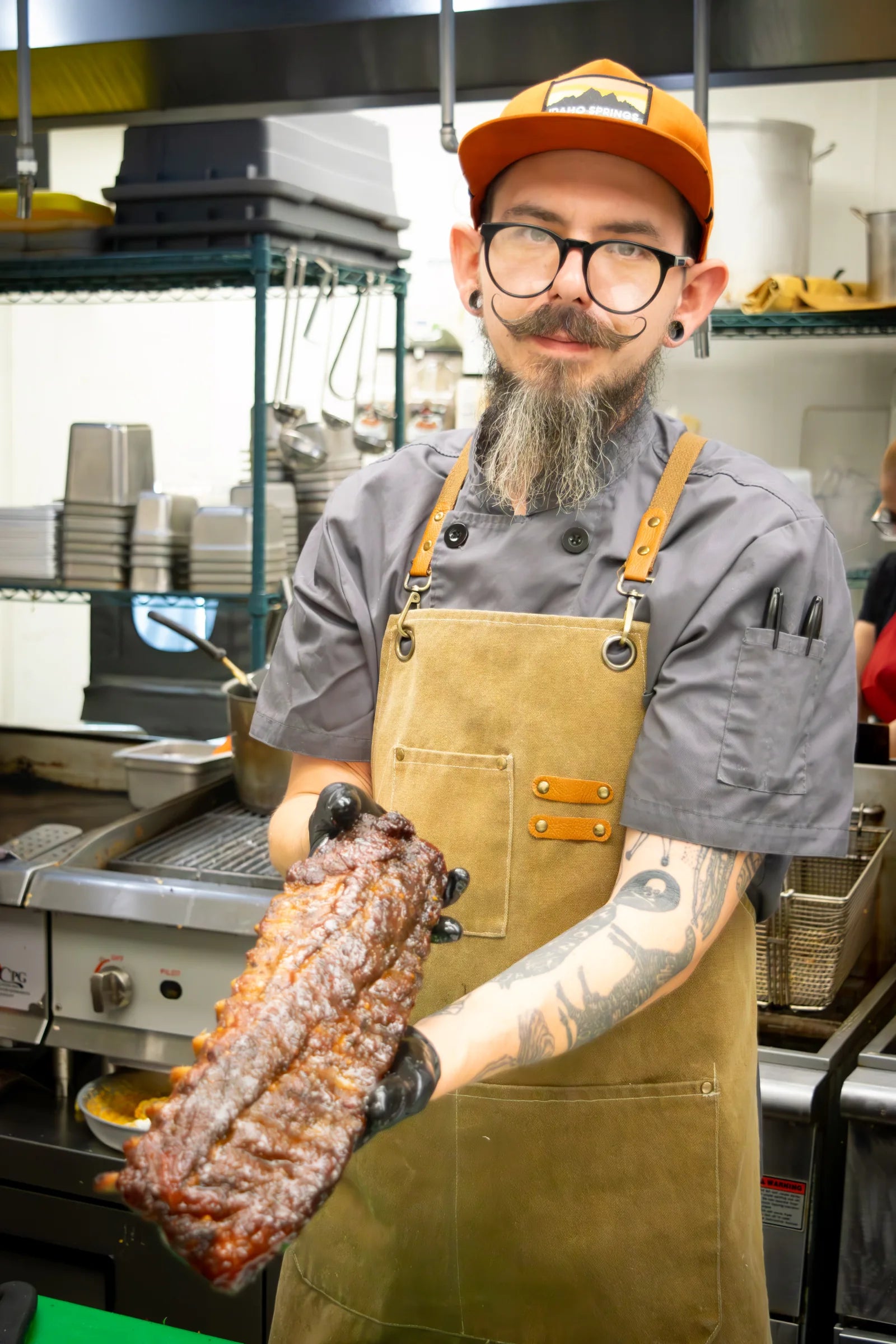 Cook wearing round glasses, an orange trucker cap, and a tan apron, holding out a full rack of smoked ribs toward the camera.
